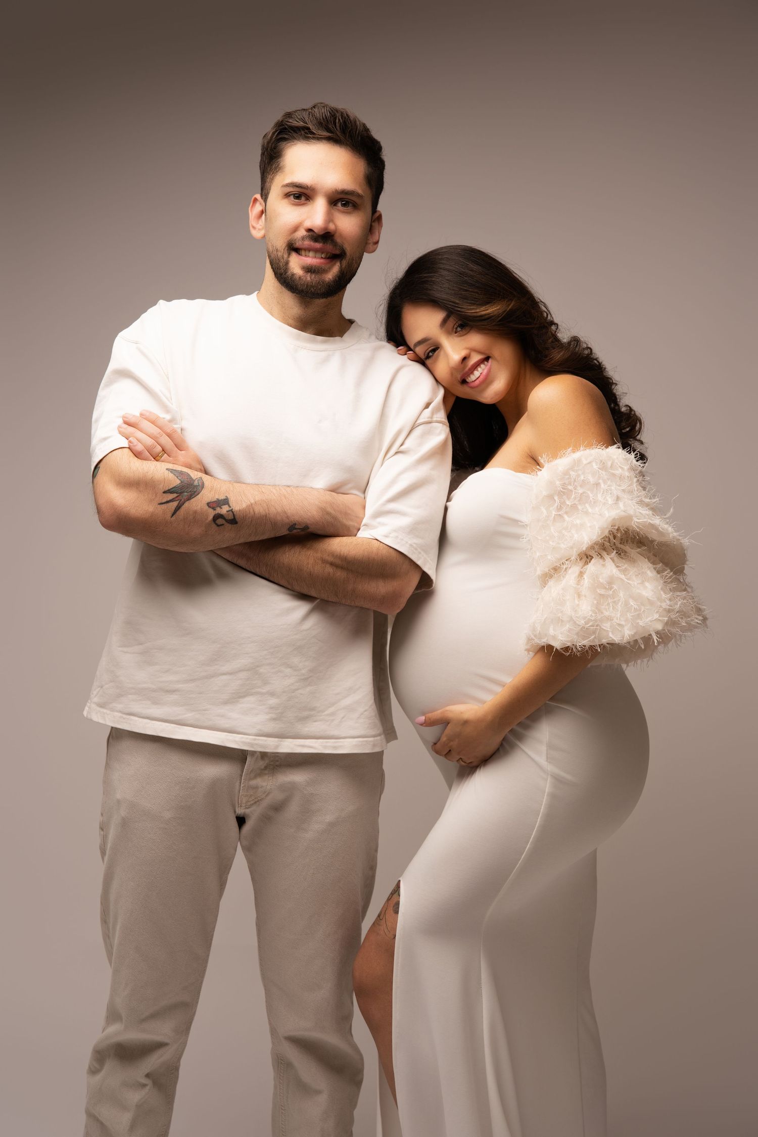 A couple poses together against neutral backdrop in white attire, embracing tenderly.