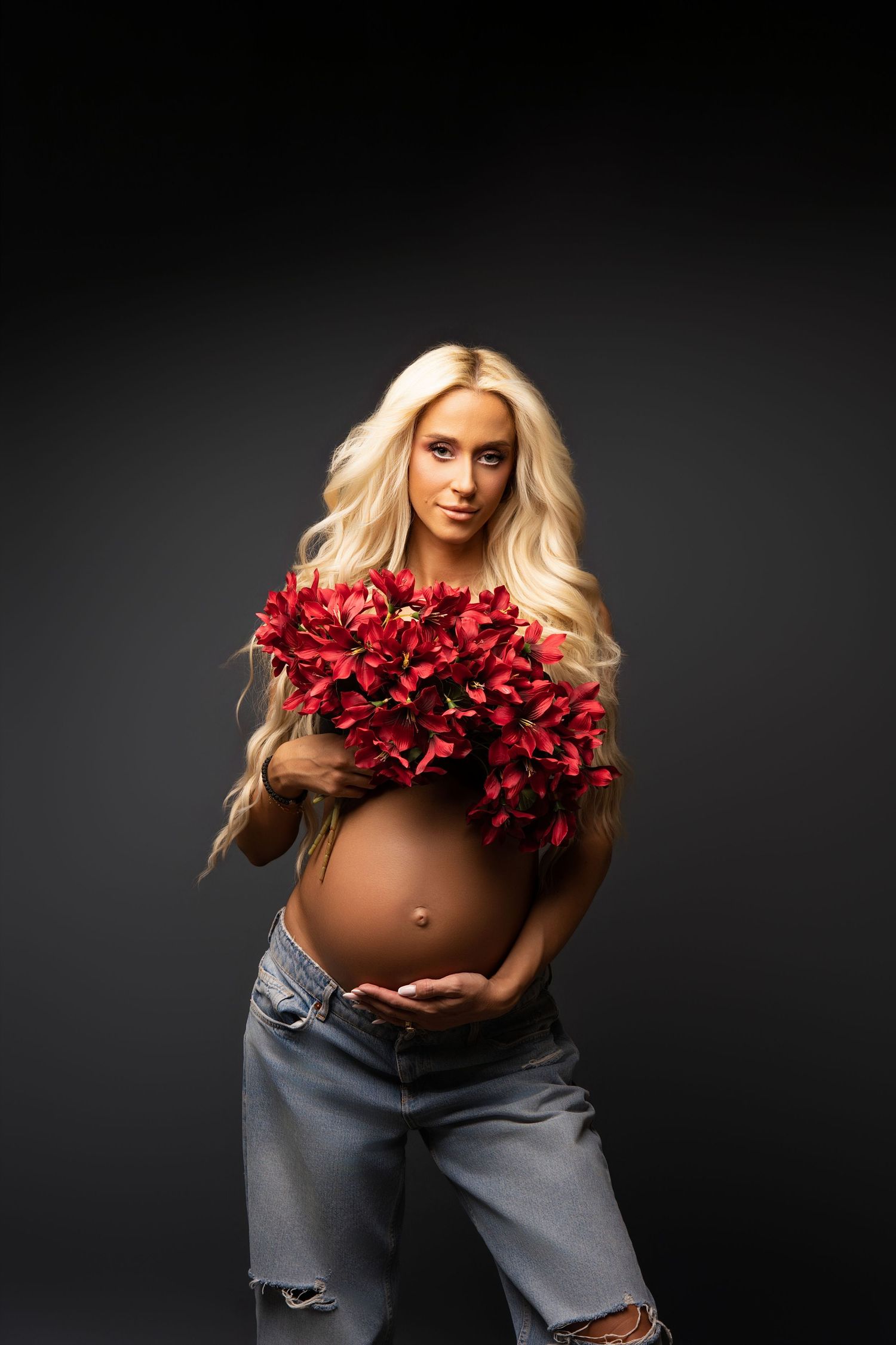 An artistic maternity photoshoot featuring red roses and denim against a dark background.