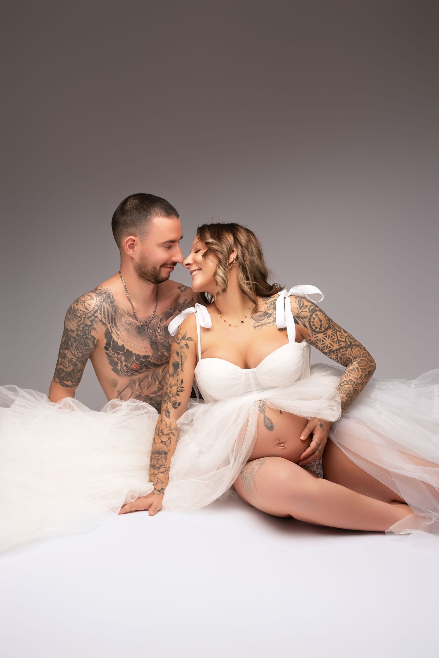 A romantic maternity photoshoot of a couple laying on white fabric with the partner showing tattoo sleeves.