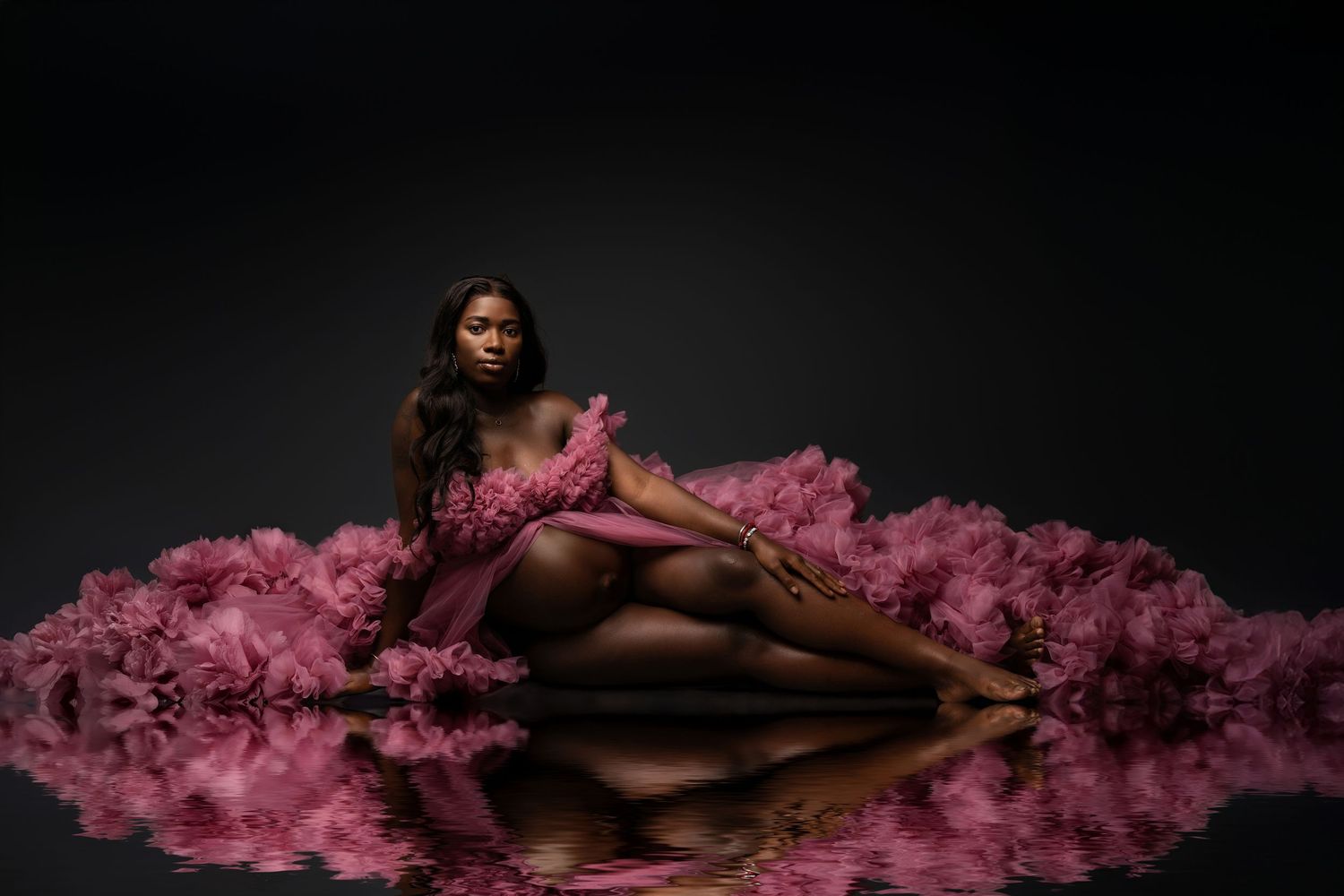 Artistic portrait of someone reclining on floor in flowing pink tulle gown against dark background with reflection.