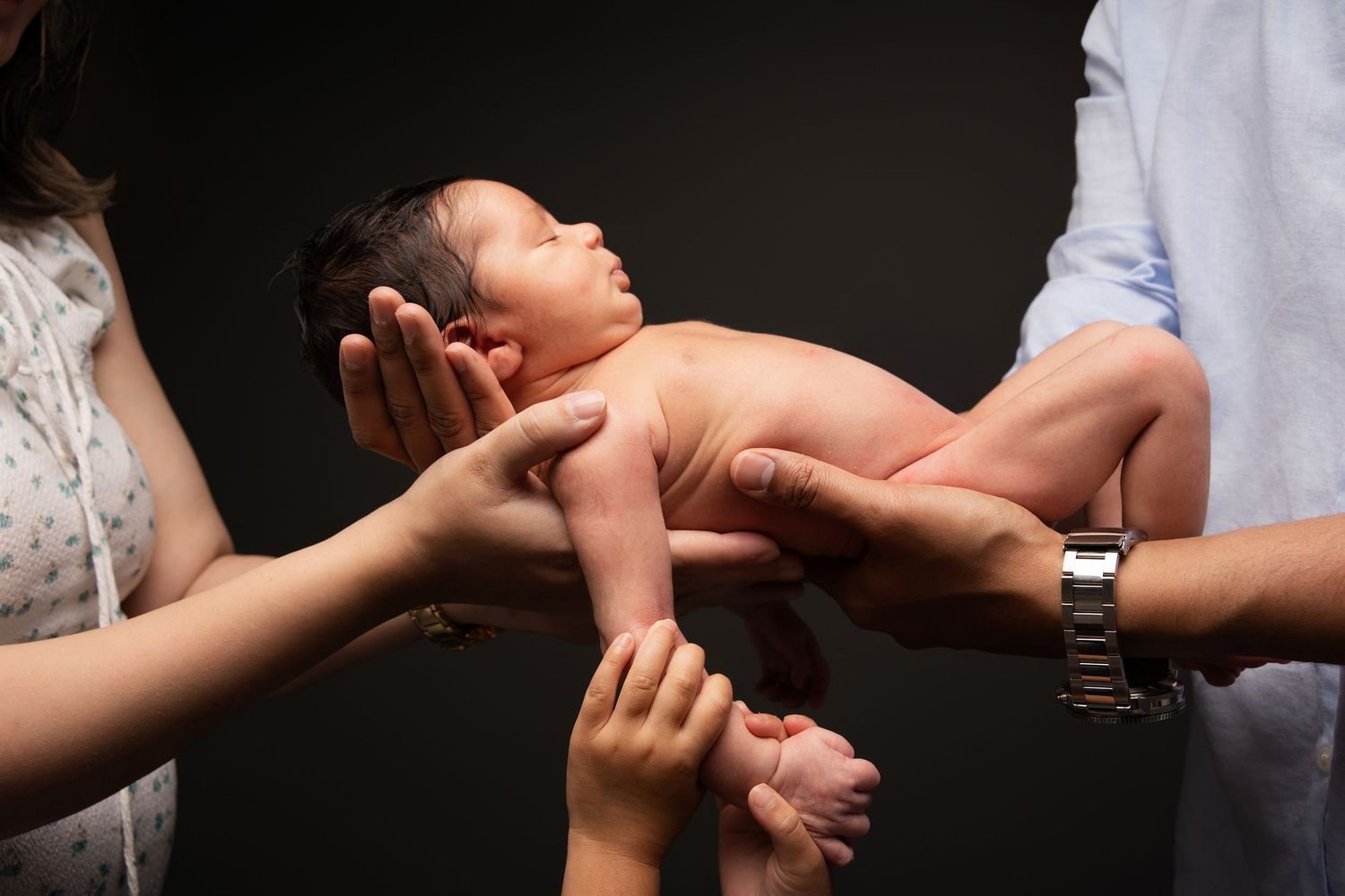 Multiple hands gently support a sleeping newborn baby against a dark background.