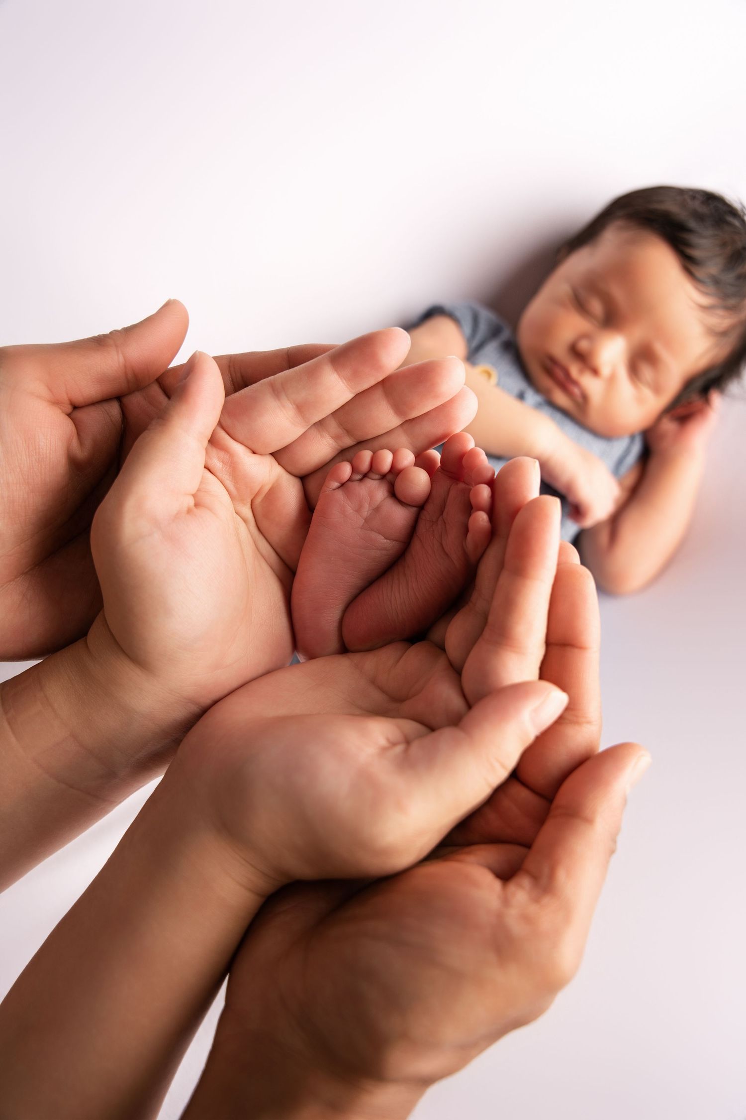 Adult hands gently cradle tiny baby feet while an infant sleeps peacefully in soft lighting.