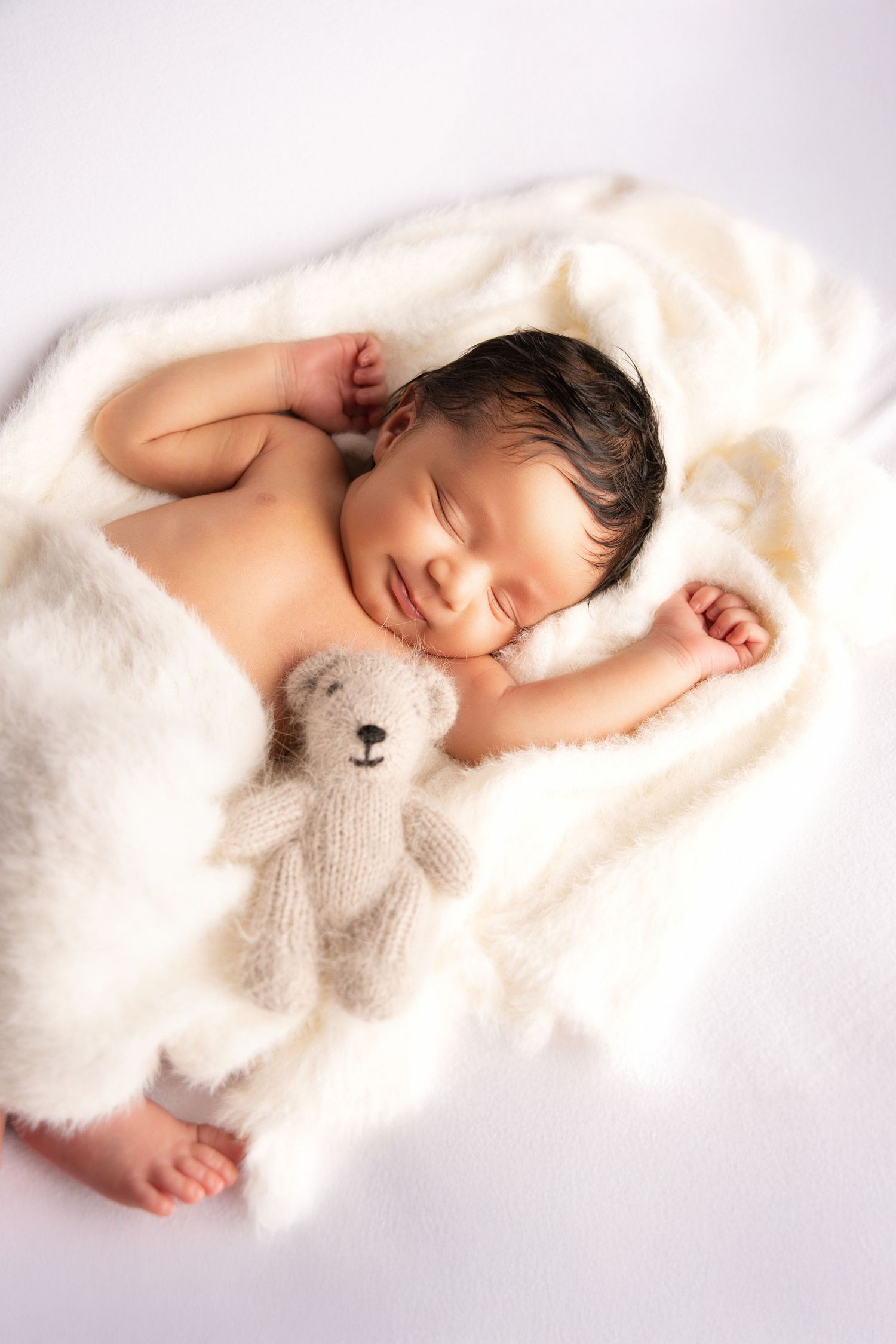 Sleeping newborn wrapped in soft white blanket cuddles with small plush teddy bear in gentle lighting.