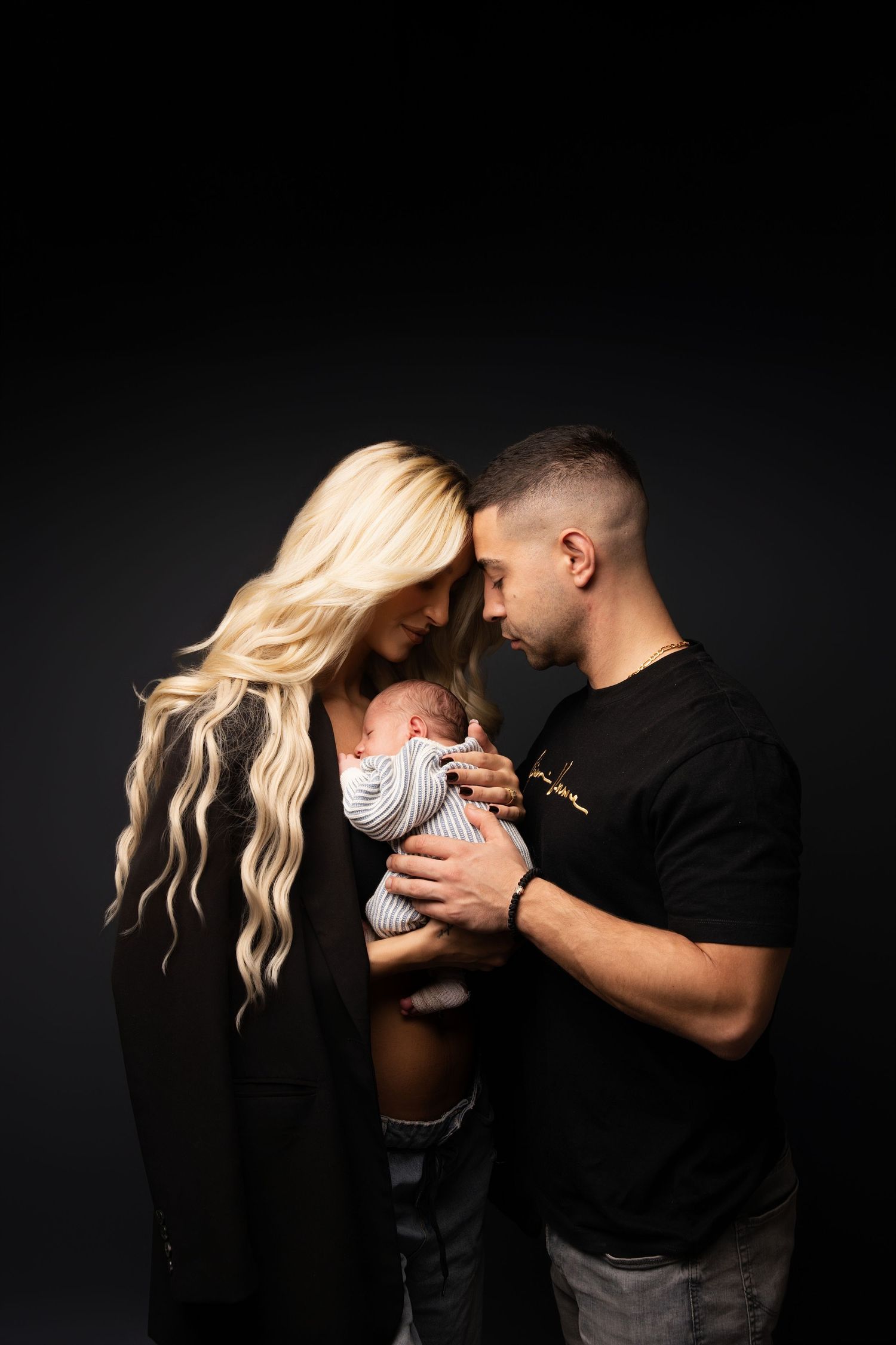 Parents sharing tender moment with their newborn baby against dark background in studio portrait setting.