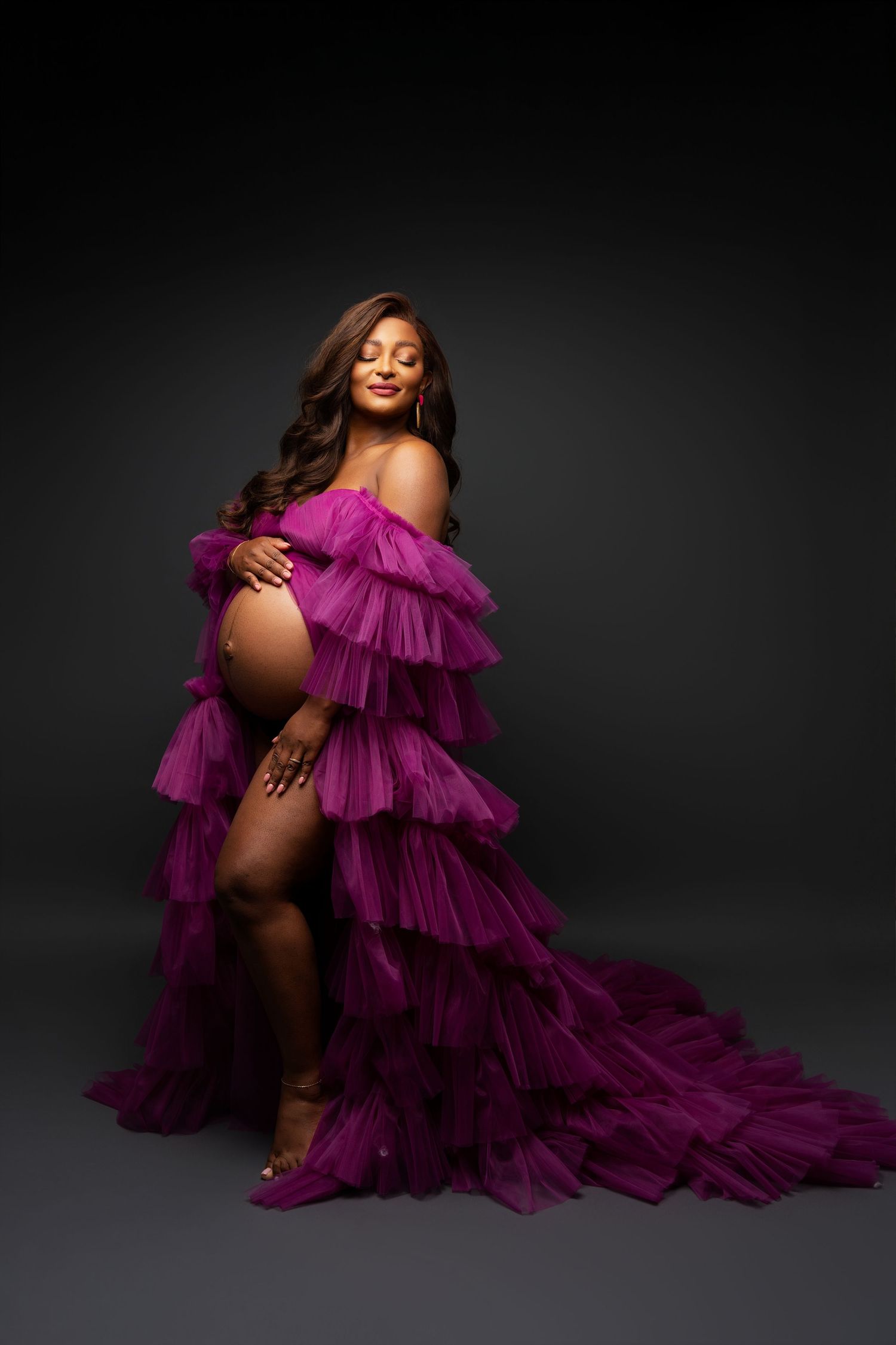 A stunning maternity portrait featuring an elegant purple tulle gown with ruffled tiers against a dark studio backdrop.