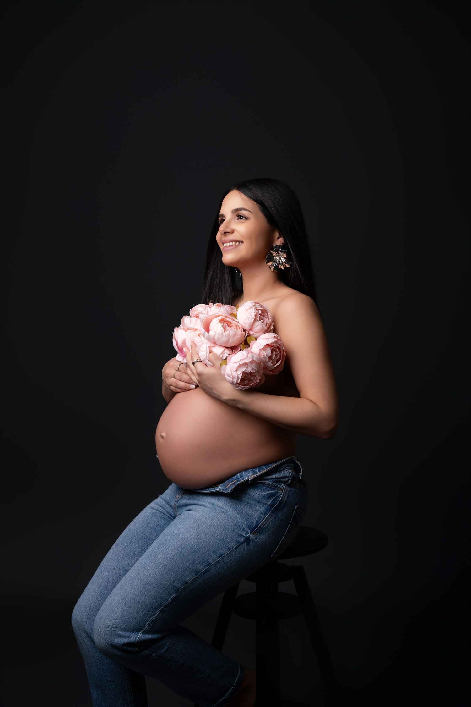 Art portrait of an expectant mother holding pink peonies against dark background in casual denim.