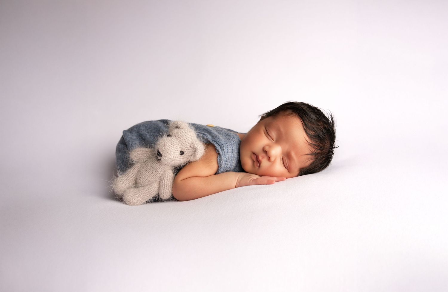 Sleeping newborn baby in light blue outfit cuddles with small white teddy bear on white background.