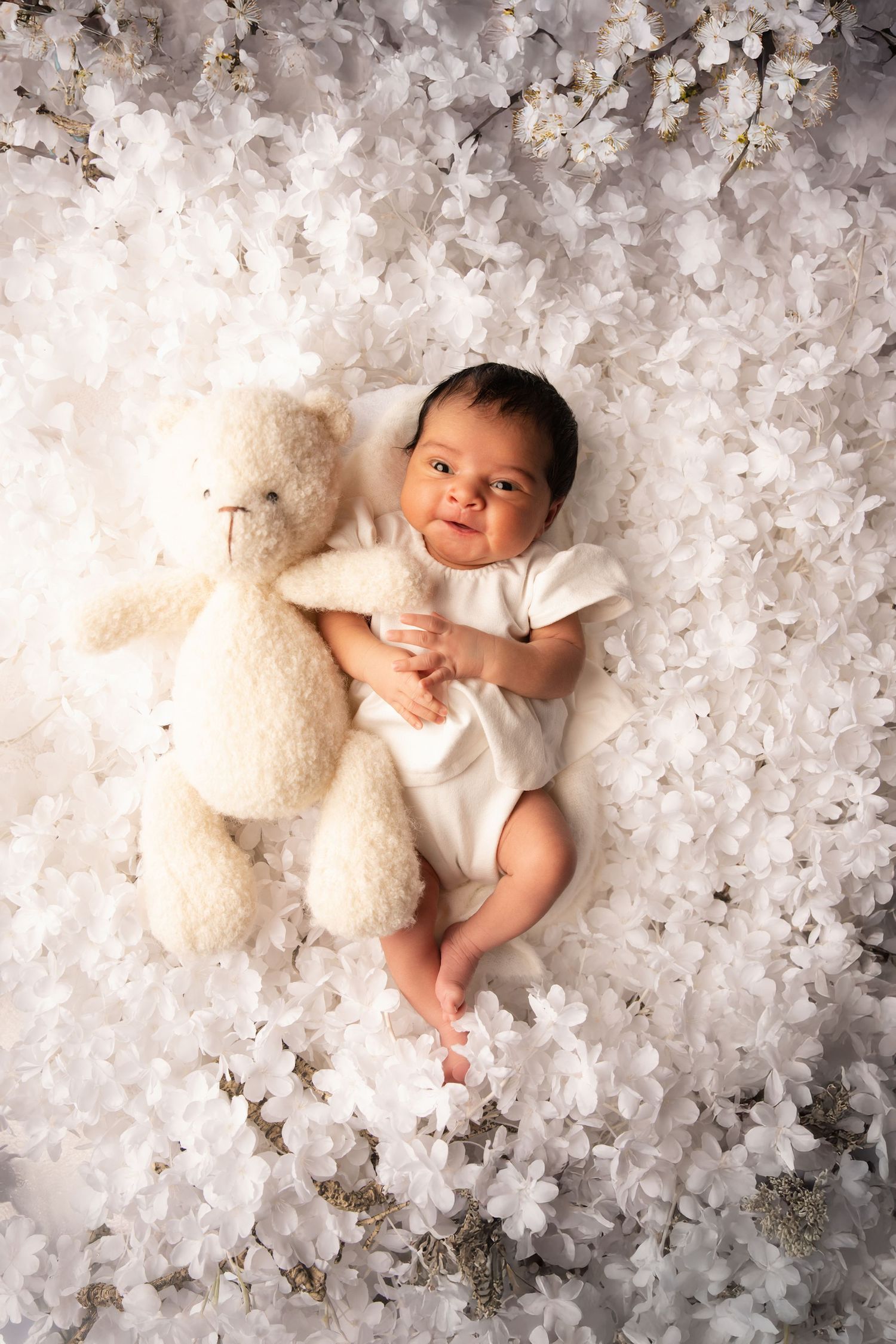 Baby in white onesie lying on soft white floral backdrop with teddy bear in professional newborn photography session.