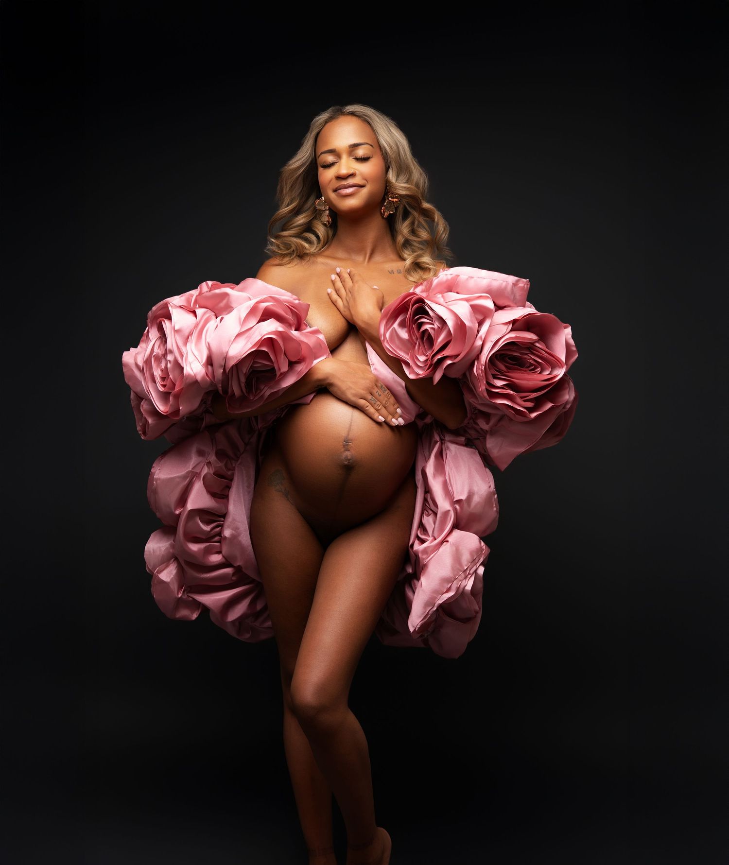 A maternity photo featuring a nude figure draped in large pink rose-like fabric against a dark background.