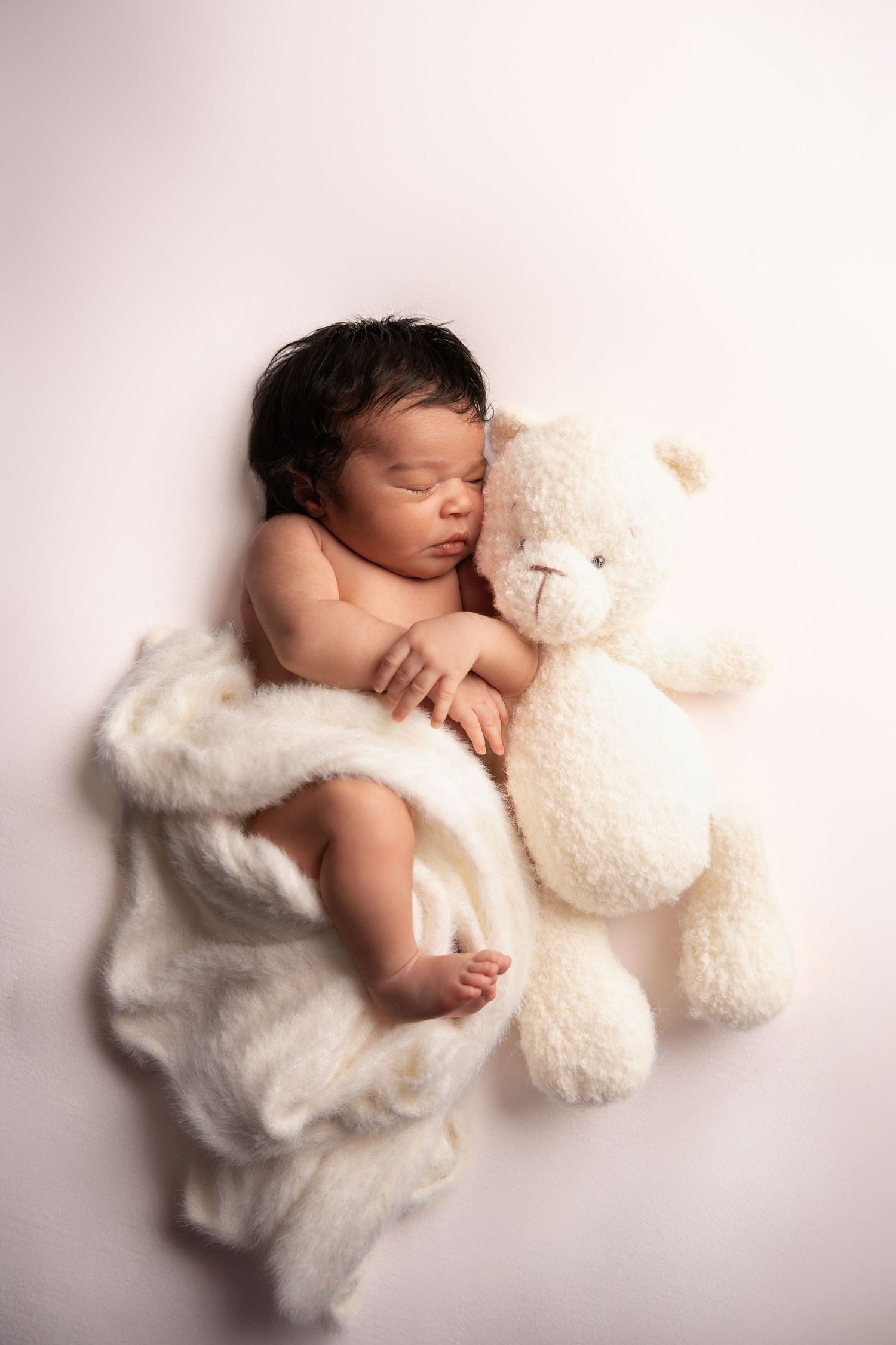 A sleeping newborn baby snuggles with a plush white teddy bear on a soft white background.