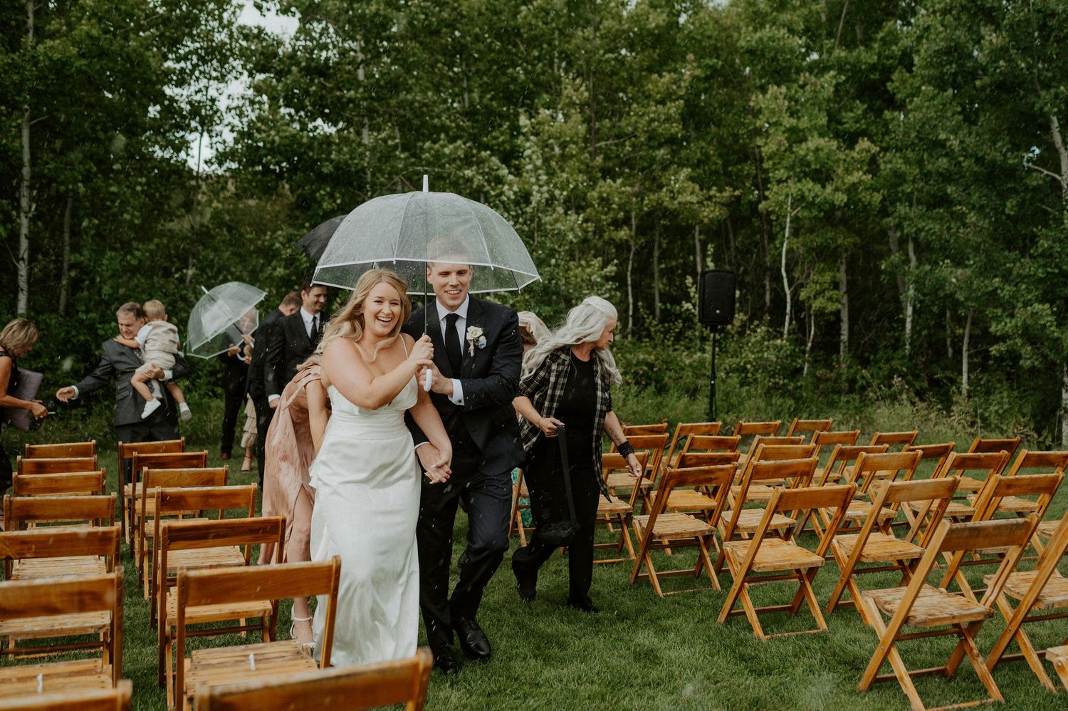 Wedding party holding umbrellas running past wooden chairs at an outdoor ceremony during heavy rain in a forest setting.