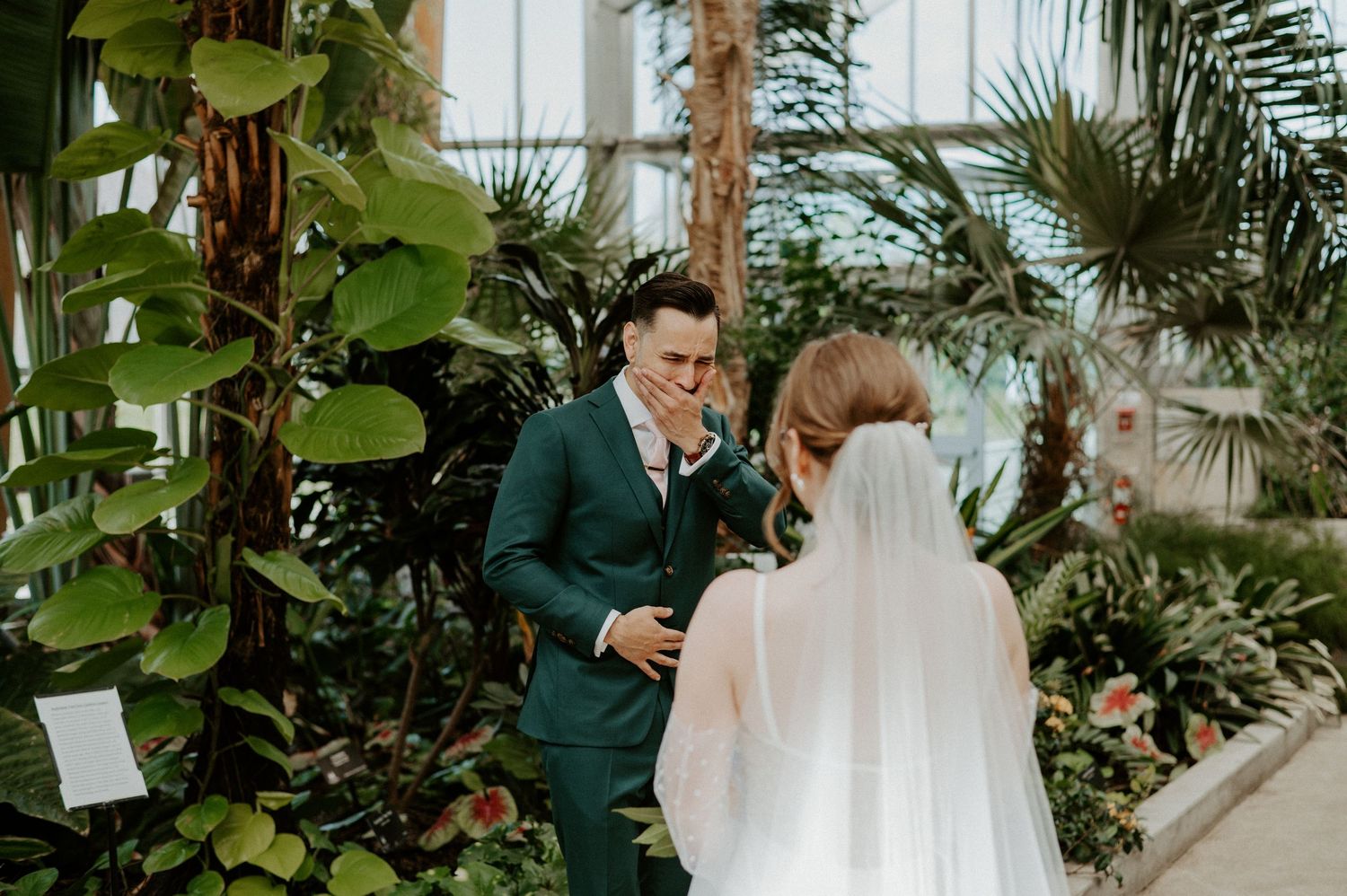 Wedding couple shares an intimate moment surrounded by lush tropical plants and palm trees in a greenhouse setting.