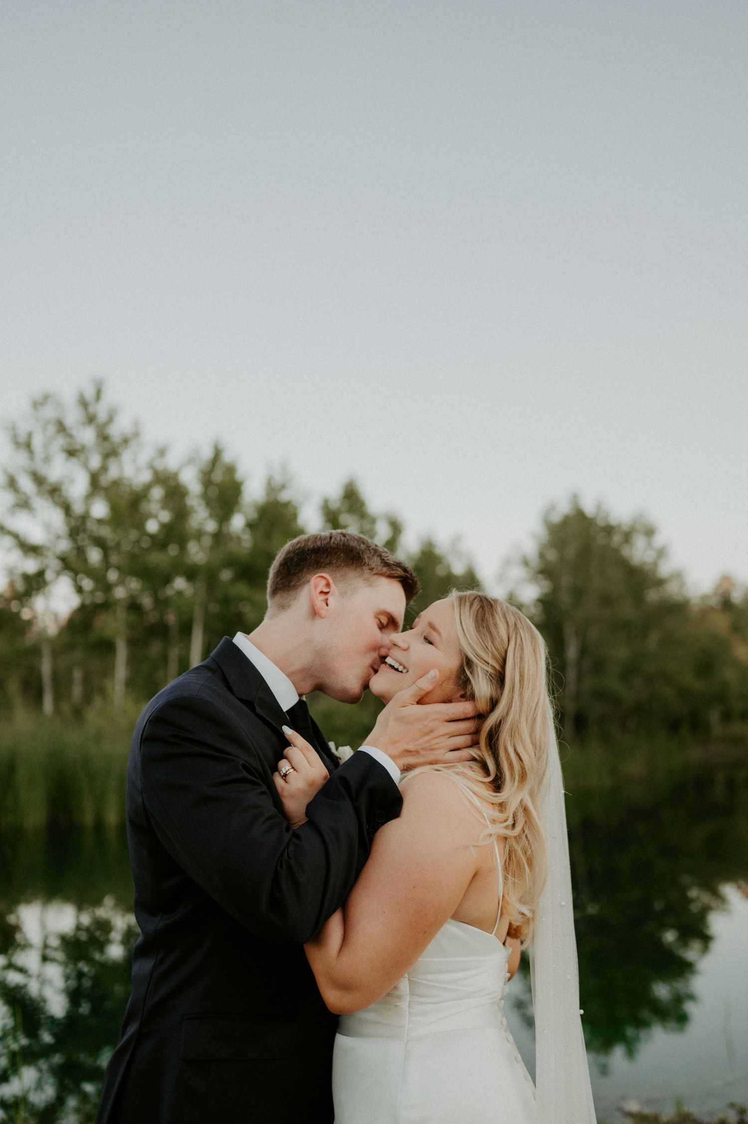 Romantic newlywed couple sharing an intimate moment by a serene lake at sunset with lush greenery in the background.