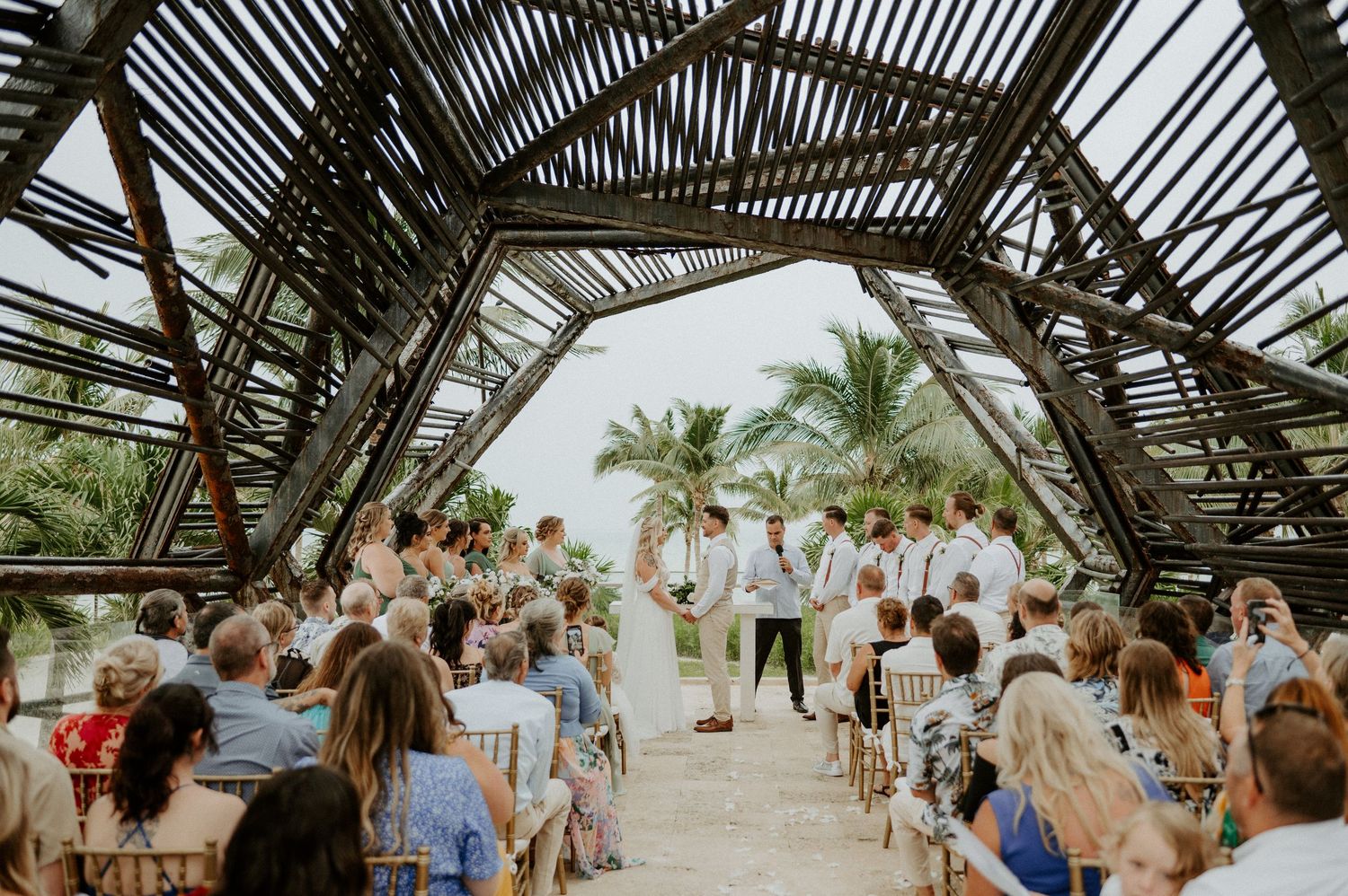 A wedding ceremony takes place under a wooden lattice structure with palm trees and ocean views in the background.