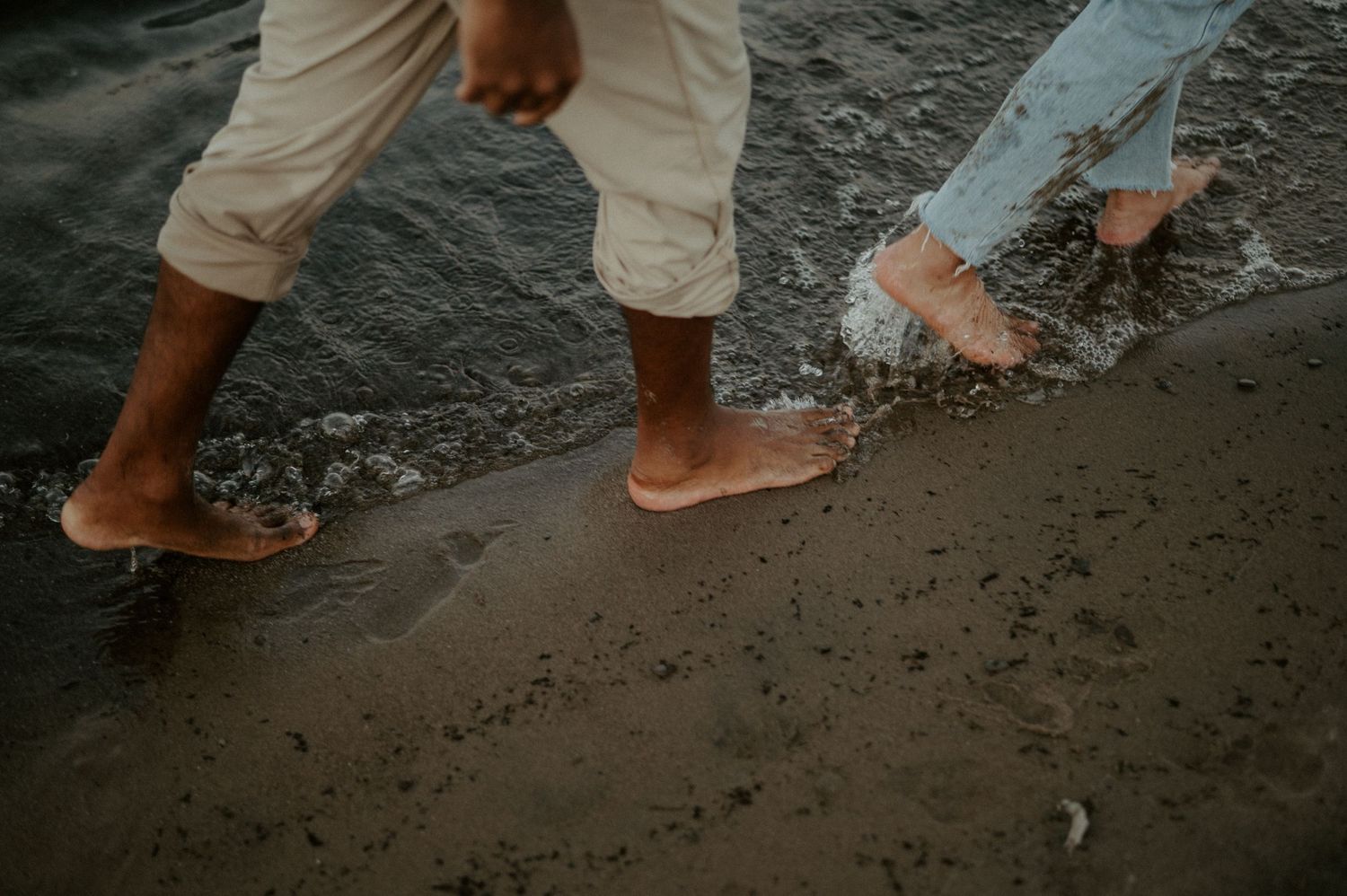 Two barefoot people walking on wet sandy beach together with their feet partially submerged in shallow water.