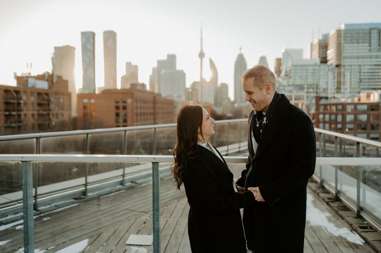 Two people stand on a rooftop balcony in Toronto at sunset with the CN Tower visible in the skyline.