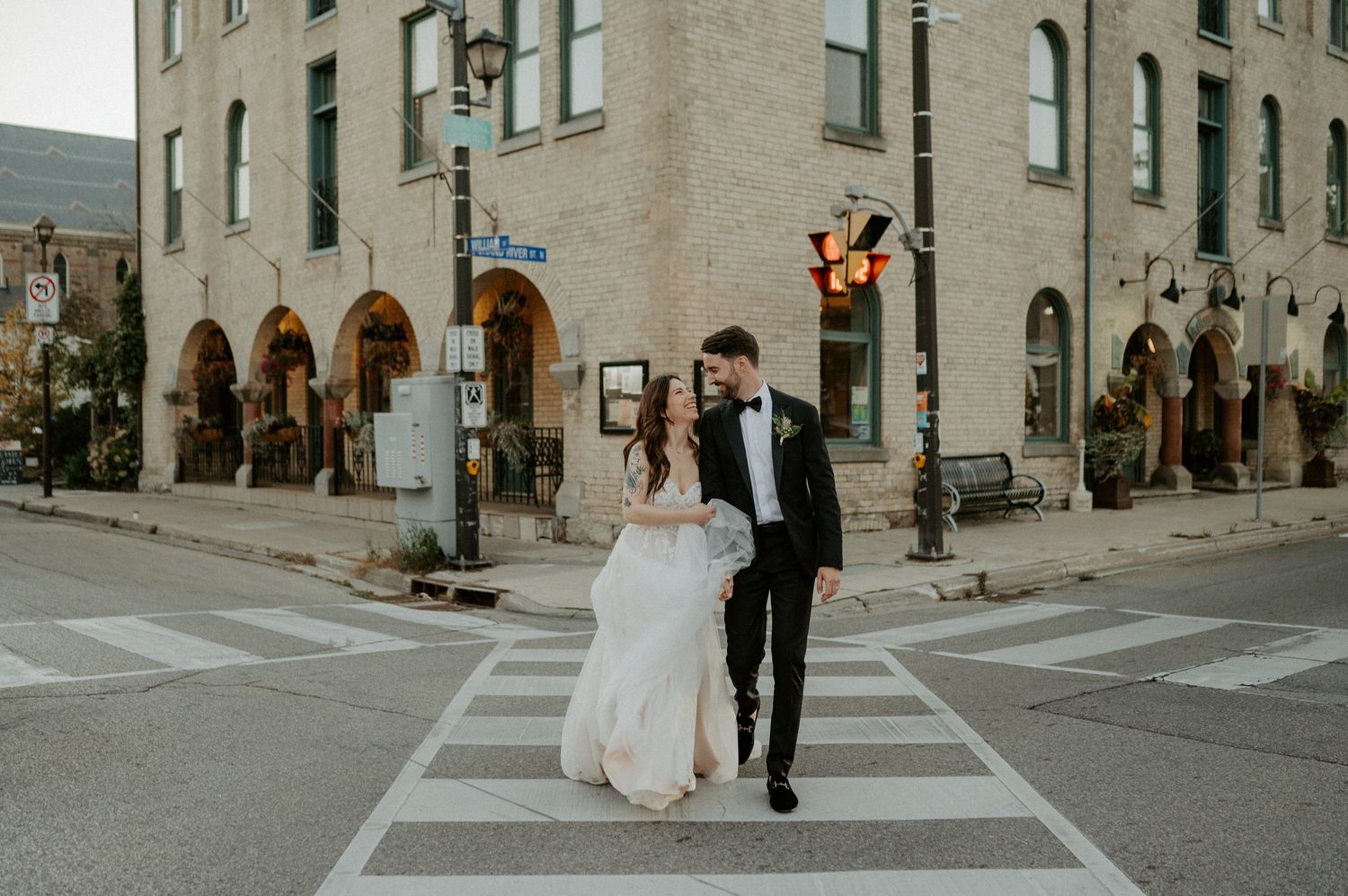 A bride and groom runs across a crosswalk in front of a historic brick building on a city corner.