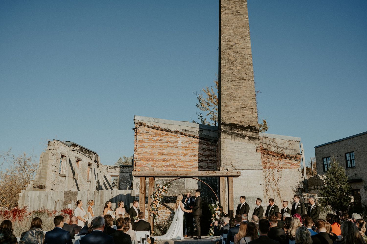 An outdoor wedding ceremony takes place at a rustic brick venue with an industrial chimney stack against a blue sky.