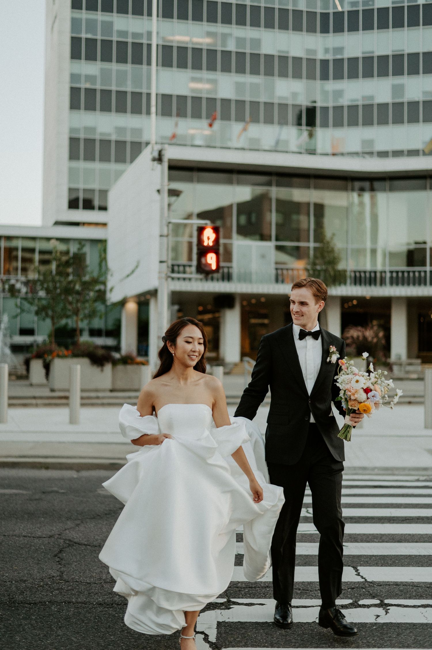 Wedding couple holding hands while crossing an urban street intersection with modern office building in background.
