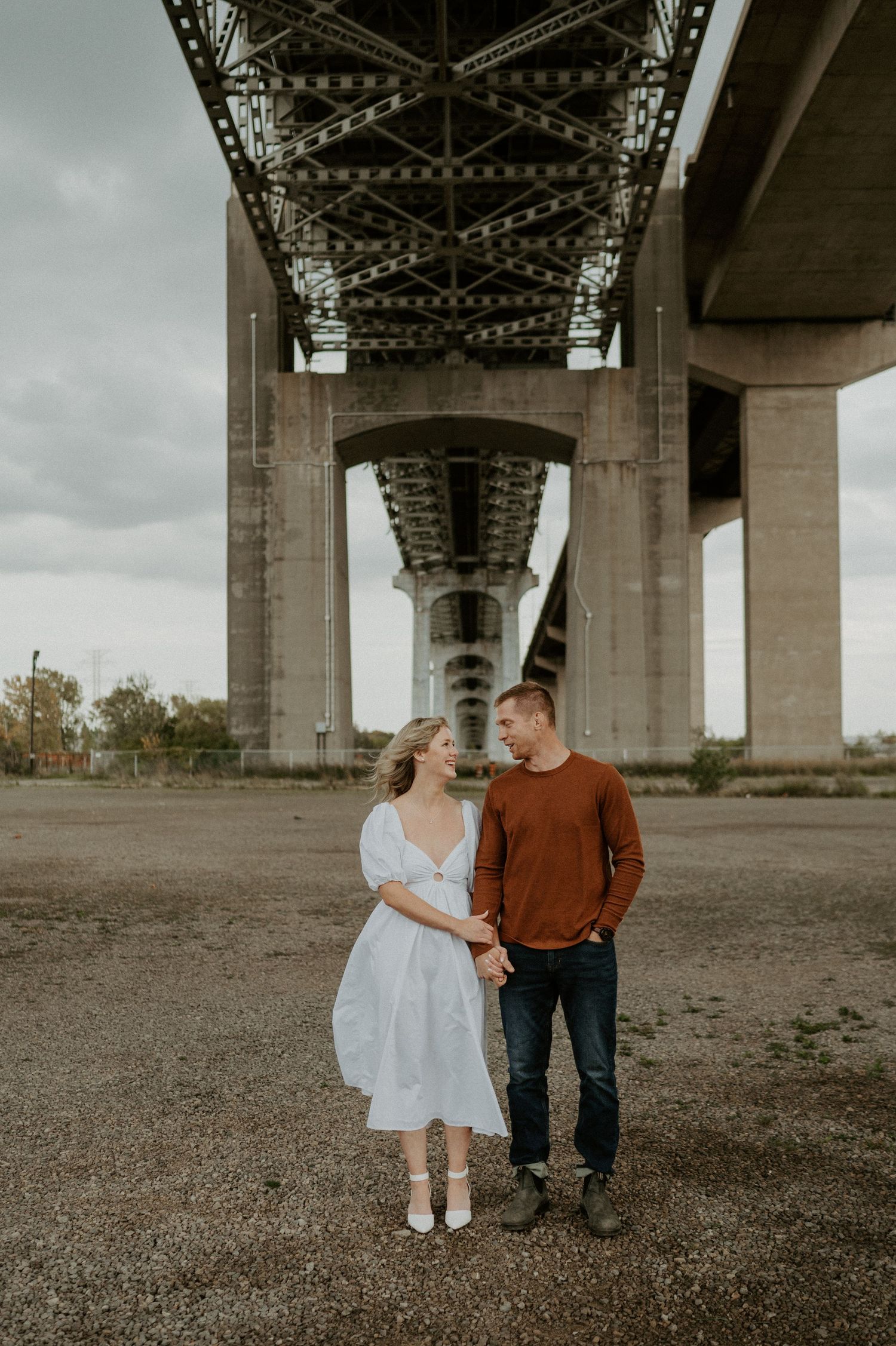 Couple standing beneath large concrete bridge on overcast day, romantic engagement style photoshoot.