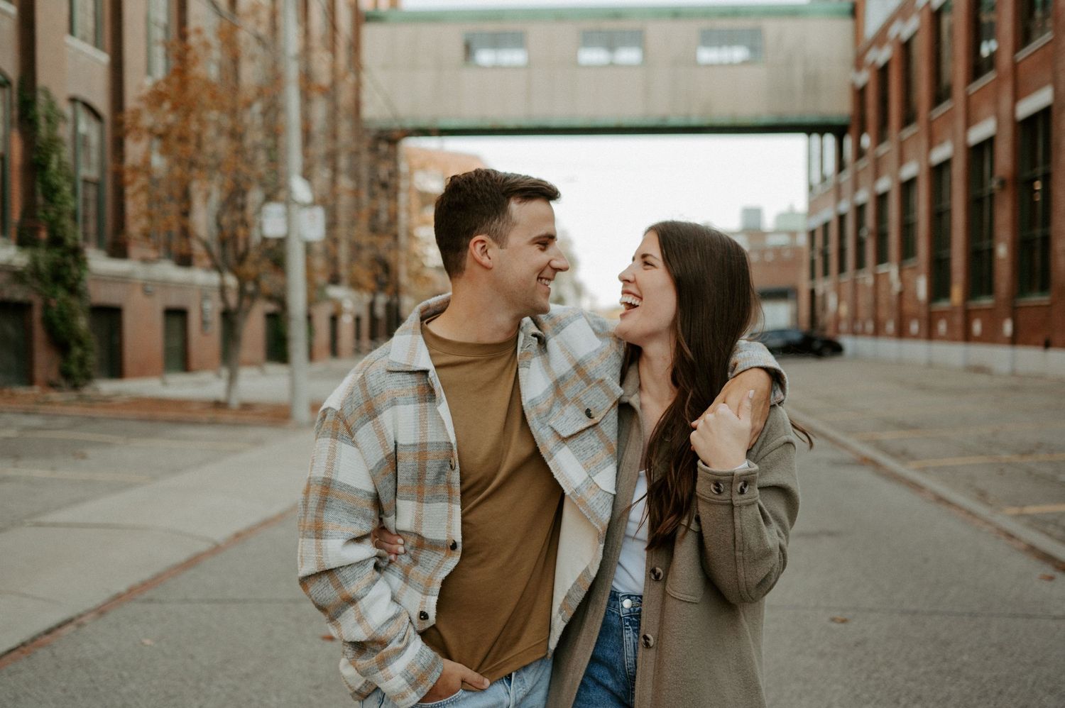 A couple shares a joyful laugh together on an urban street with brick buildings and an overhead walkway.