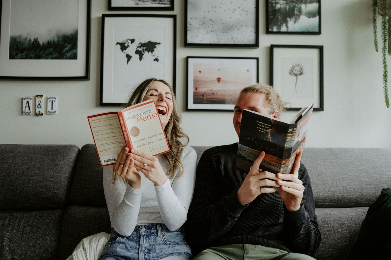 Two people sit on a gray couch laughing while holding books in front of their faces with framed artwork on the wall behind them.