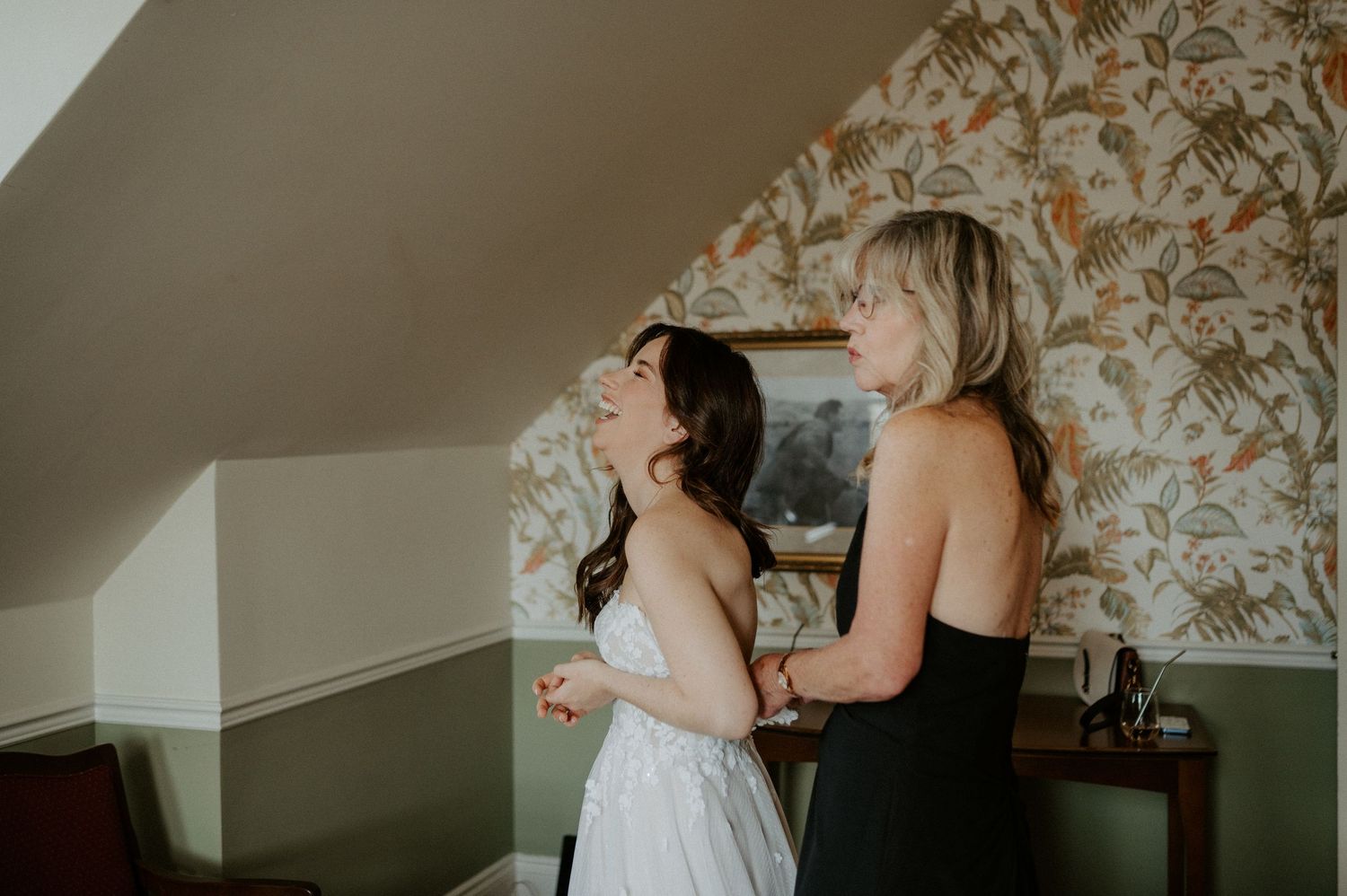 Two people in a room with floral wallpaper, one wearing a white dress while getting ready for a special occasion.