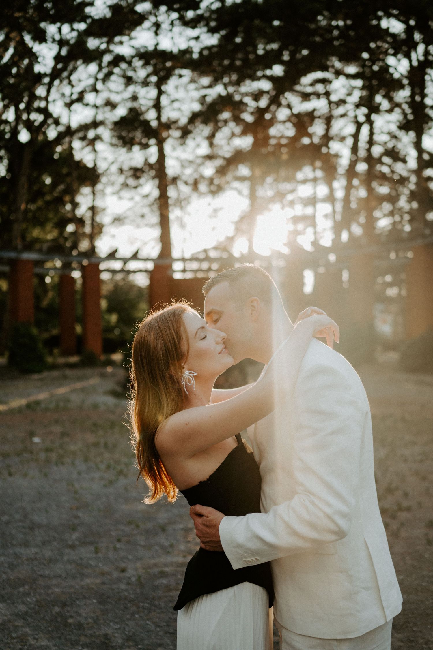 Romantic couple embraces during golden sunset in a forest, with warm sunlight filtering through pine trees.