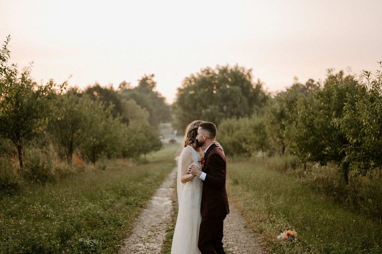 A couple shares a romantic moment on a gravel path surrounded by lush greenery at dusk.
