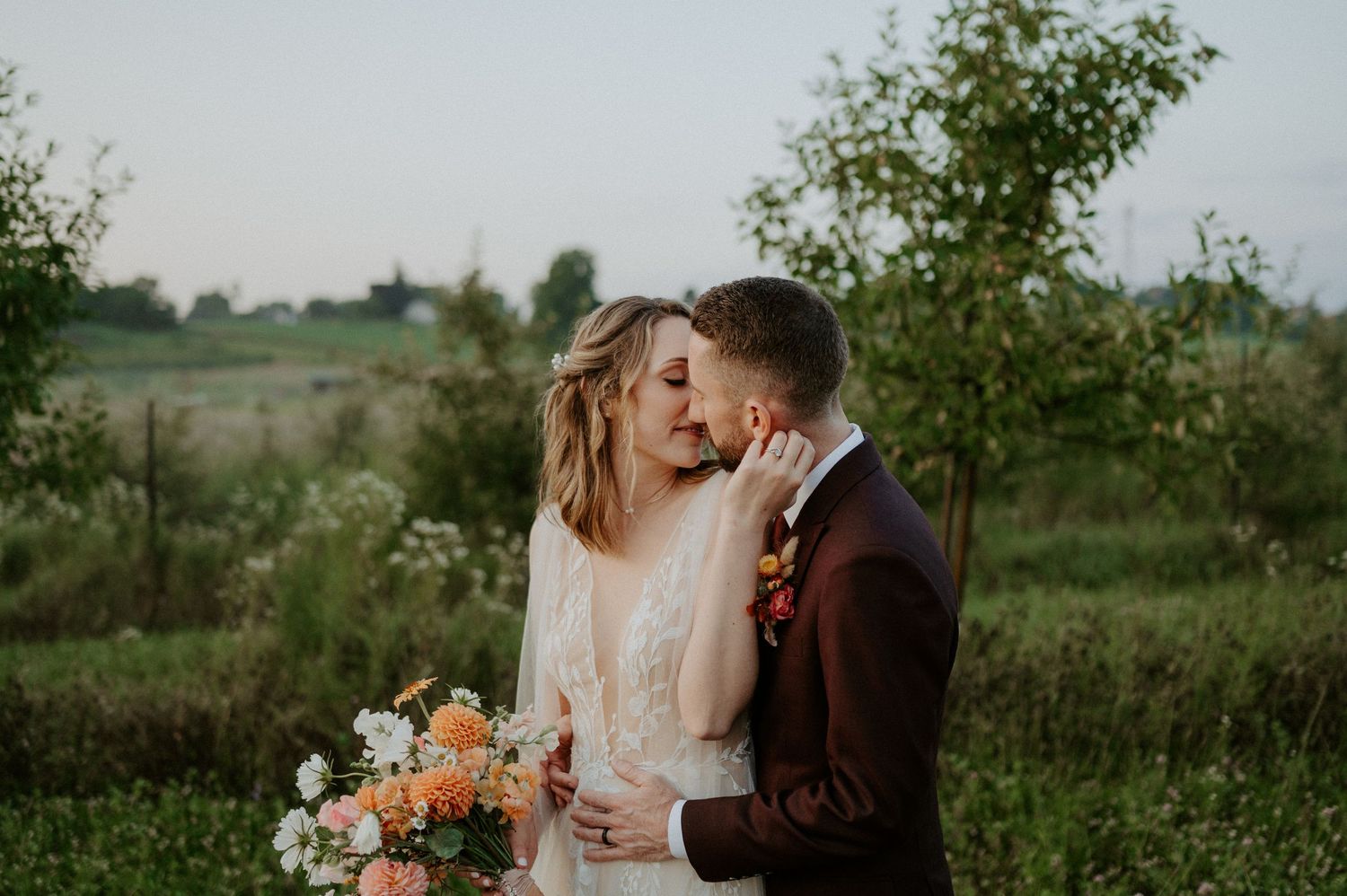 A loving couple embraces outdoors with a peach-colored floral bouquet in a rustic countryside setting at sunset.