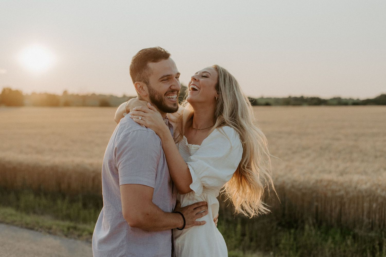A loving couple laughs together in a sunlit wheat field at golden hour.