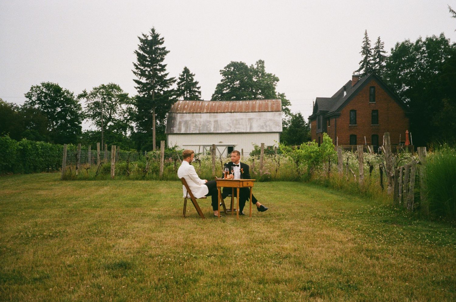 35mm film image of a couple sitting at a rustic wooden table in a backyard with a white barn and brick house in the background.