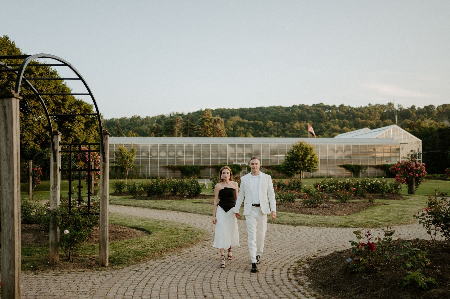 A couple walks along a curved garden path with greenhouses visible in the background on a sunny day.
