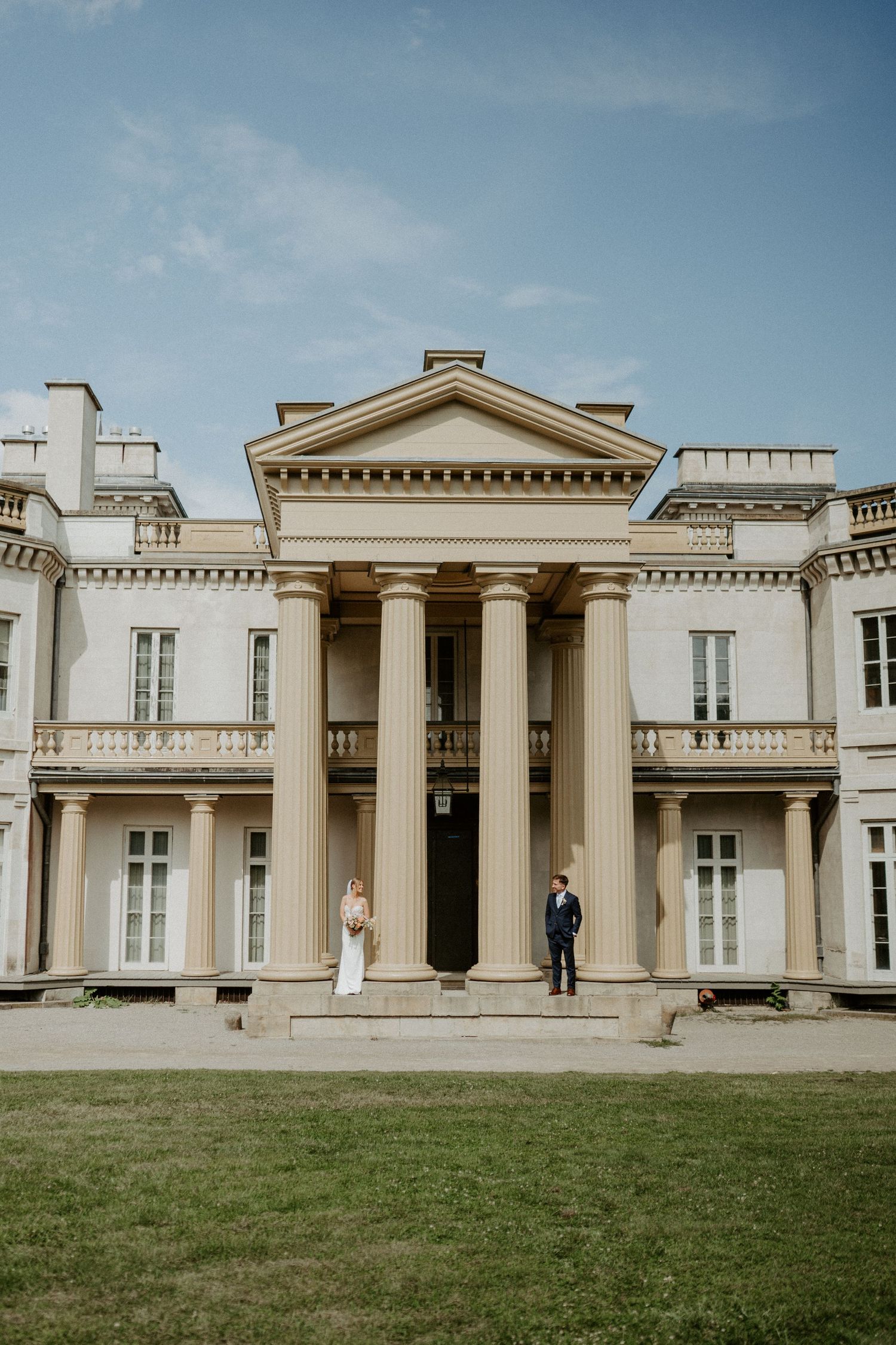A couple stands on the steps of a grand neoclassical mansion with white columns and symmetrical architecture.
