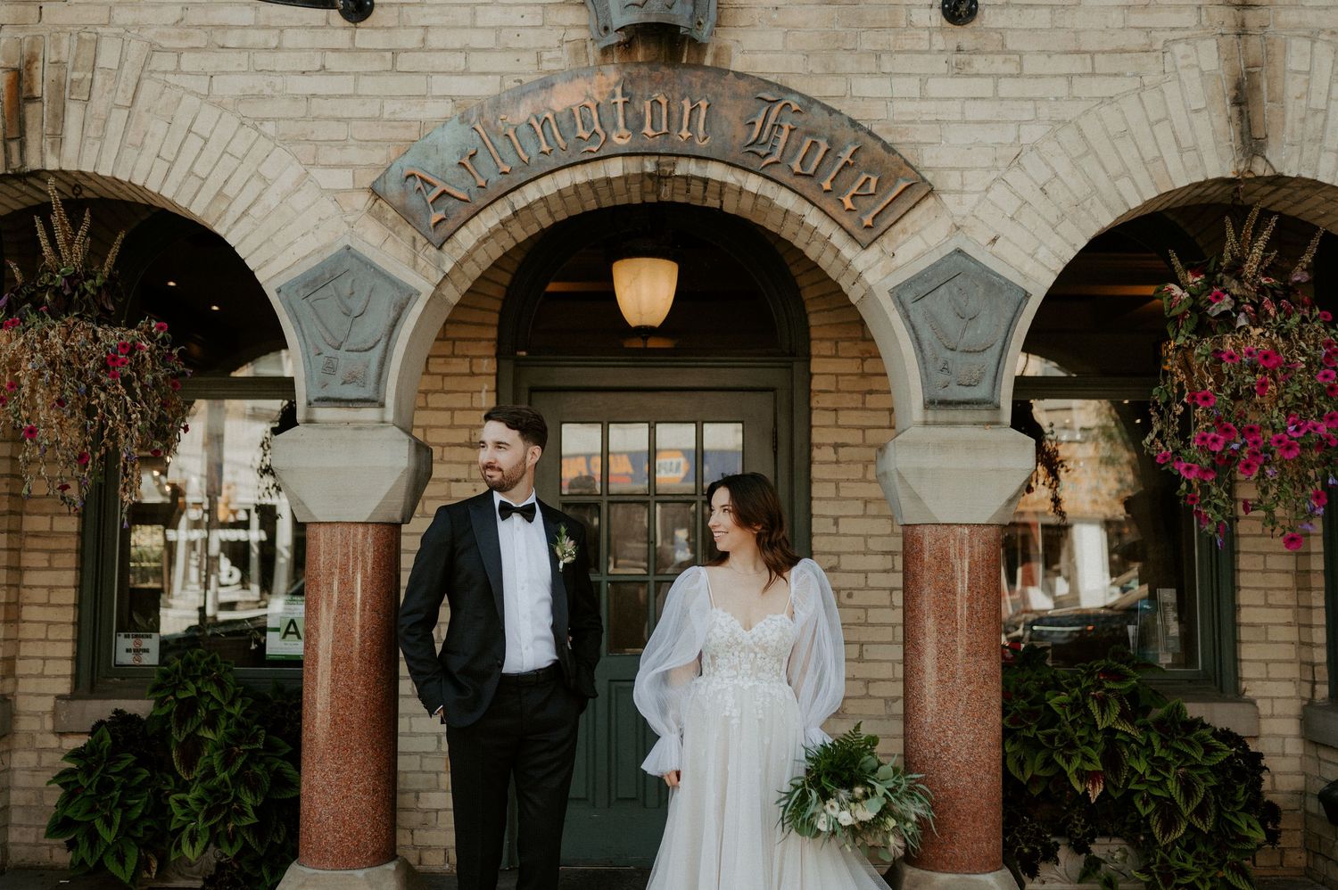 A couple stands in front of a historic brick building entrance with stone archways and hanging floral baskets.