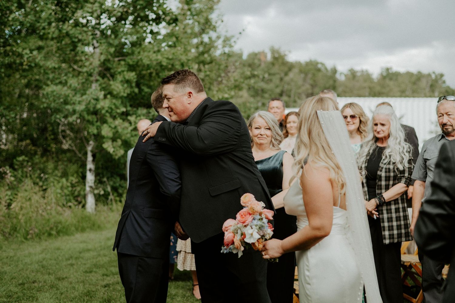An emotional outdoor wedding ceremony moment with guests gathered under cloudy skies in a green field.