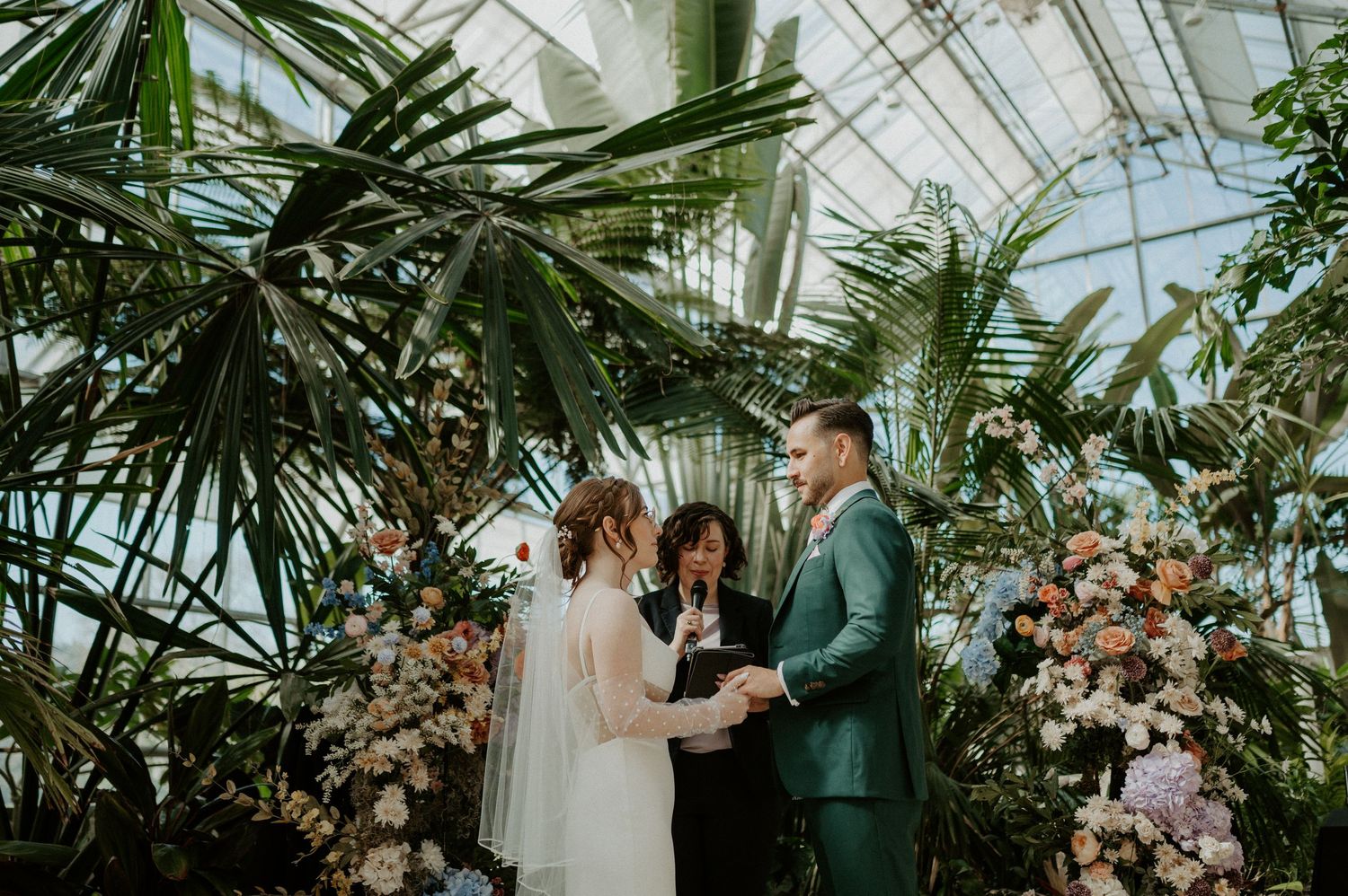 A dreamy wedding ceremony takes place inside a lush greenhouse filled with tropical plants and floral arrangements.