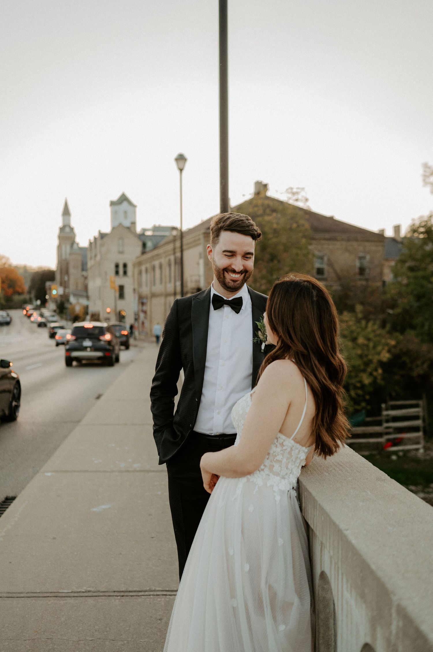A bride and groom share an intimate moment on a city bridge with a church and autumn scenery in the background.