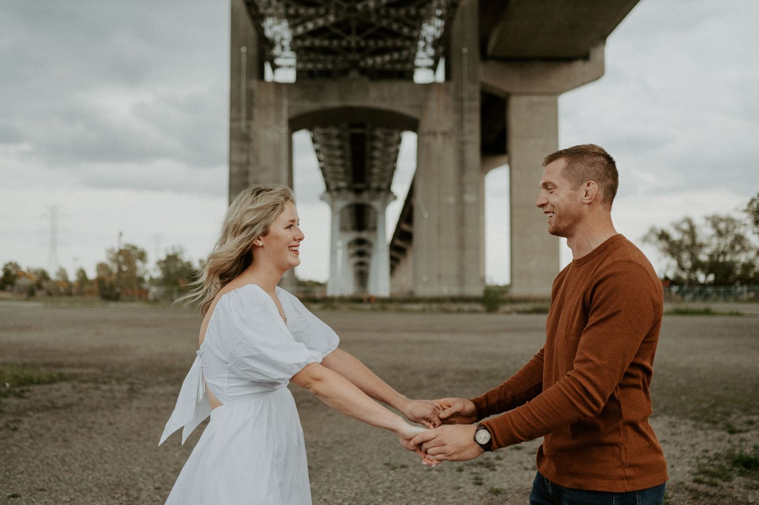 A couple shares a romantic moment under a towering bridge on an overcast day.
