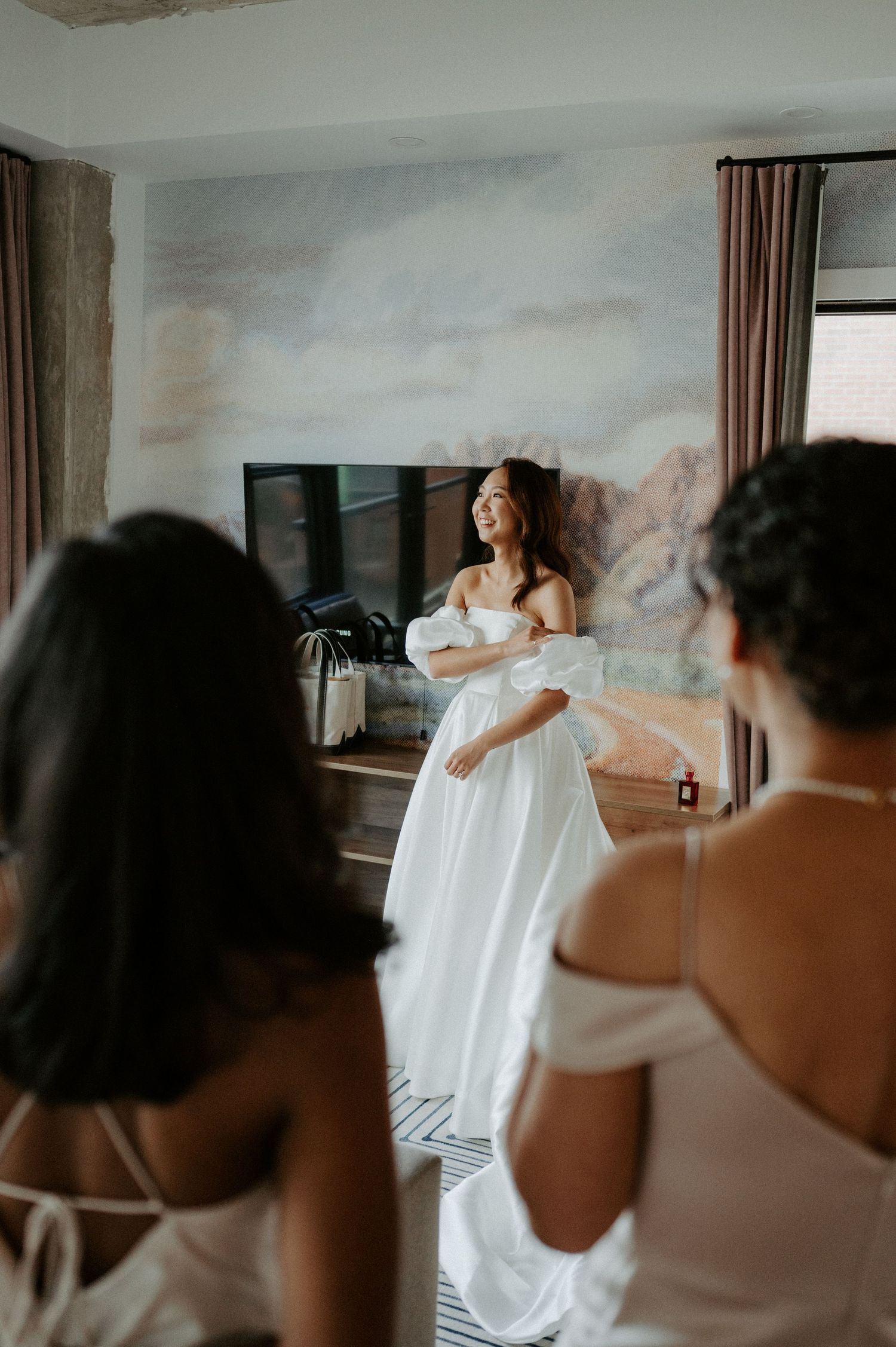 A bride in her flowing white dress stands by a window during a photography session in a room with muted wall art.