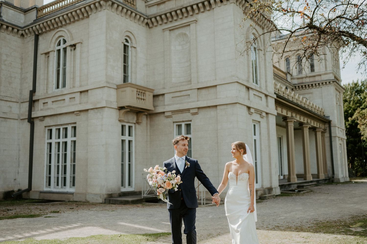 A elegant wedding couple walks hand in hand outside a grand Victorian-style mansion with white stone architecture.