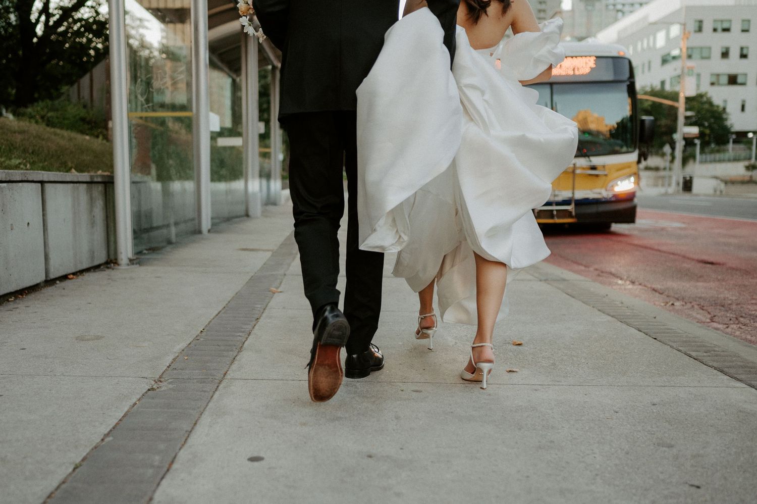 Close-up view of a couple's feet walking on sidewalk, with white wedding dress flowing and bus visible in background.