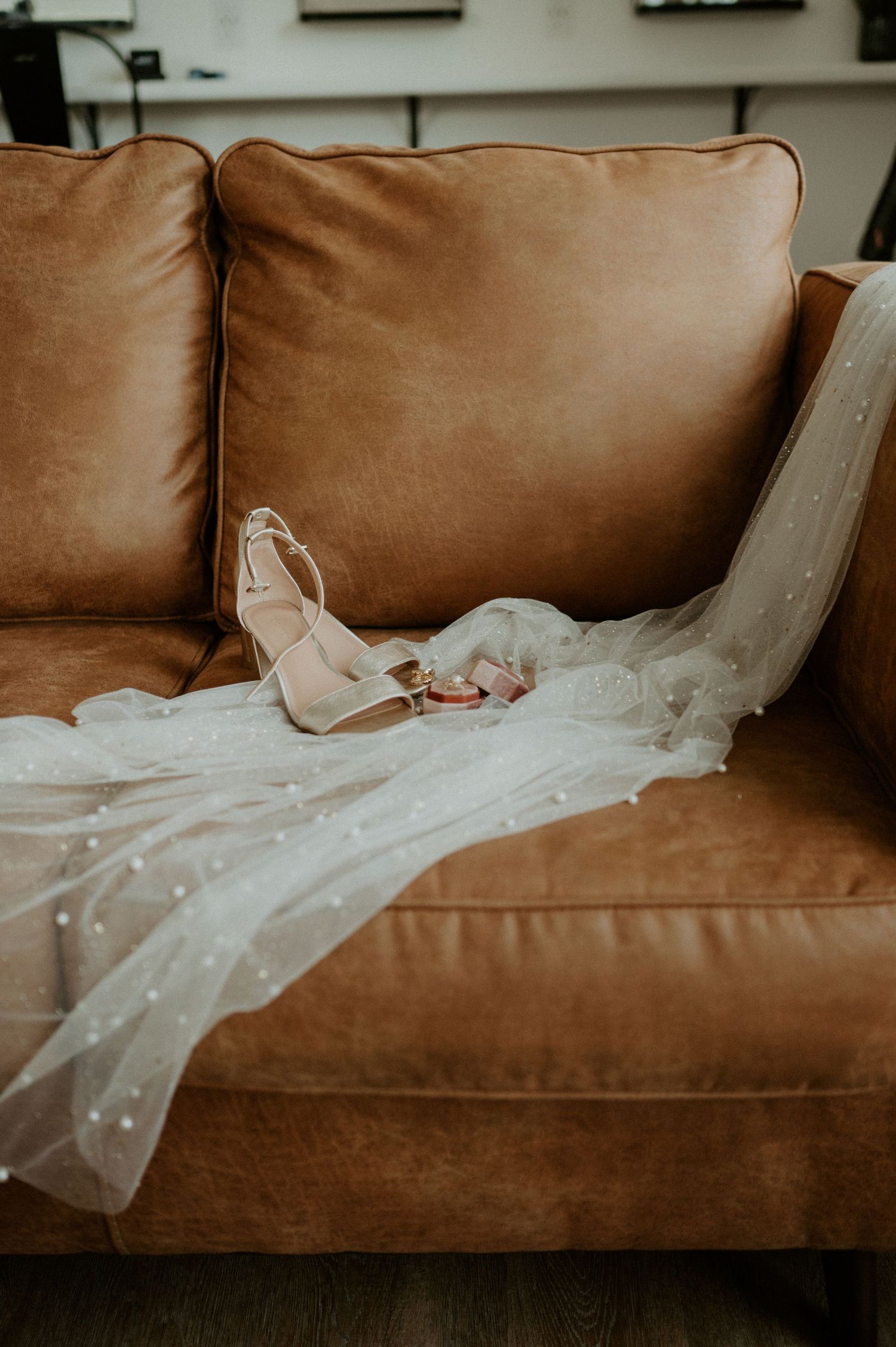 A delicate white wedding veil and heals elegantly draped across a brown leather couch.