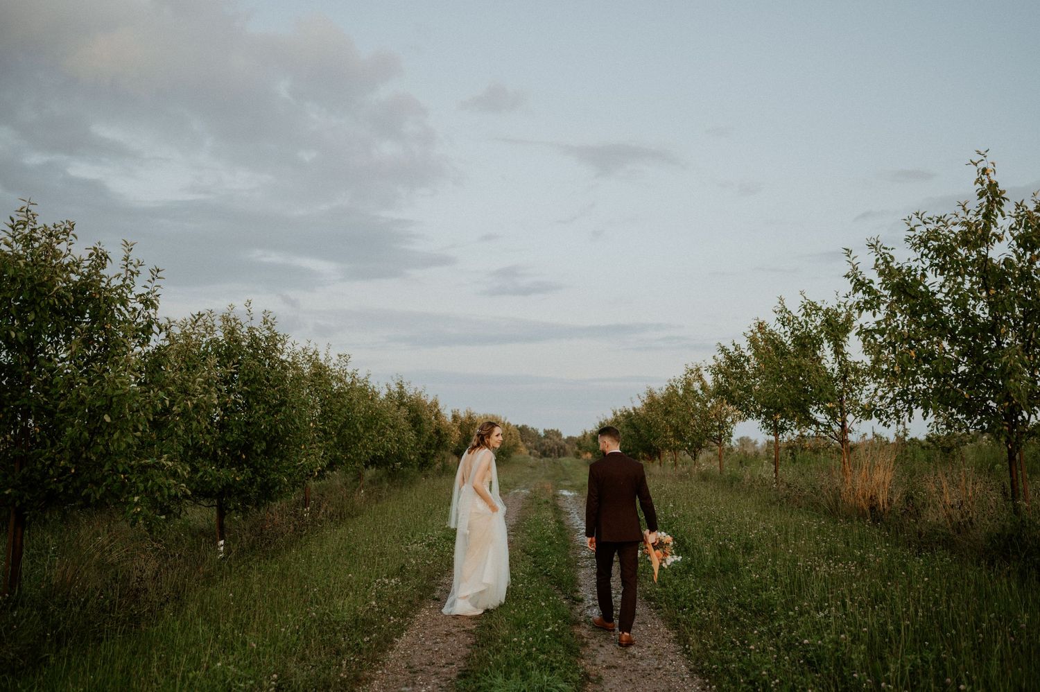 A couple walks down a grassy path between rows of apple trees at sunset holding baskets.