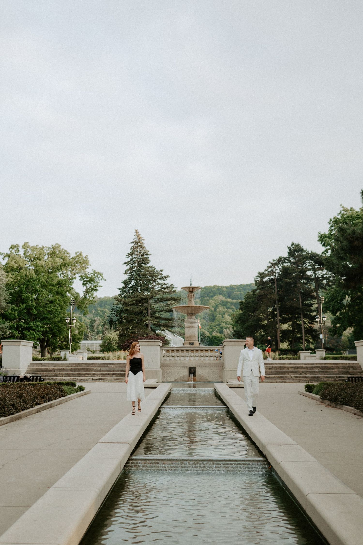 Two people in white and brown outfits stand at opposite sides of a long reflecting pool in a formal garden setting.