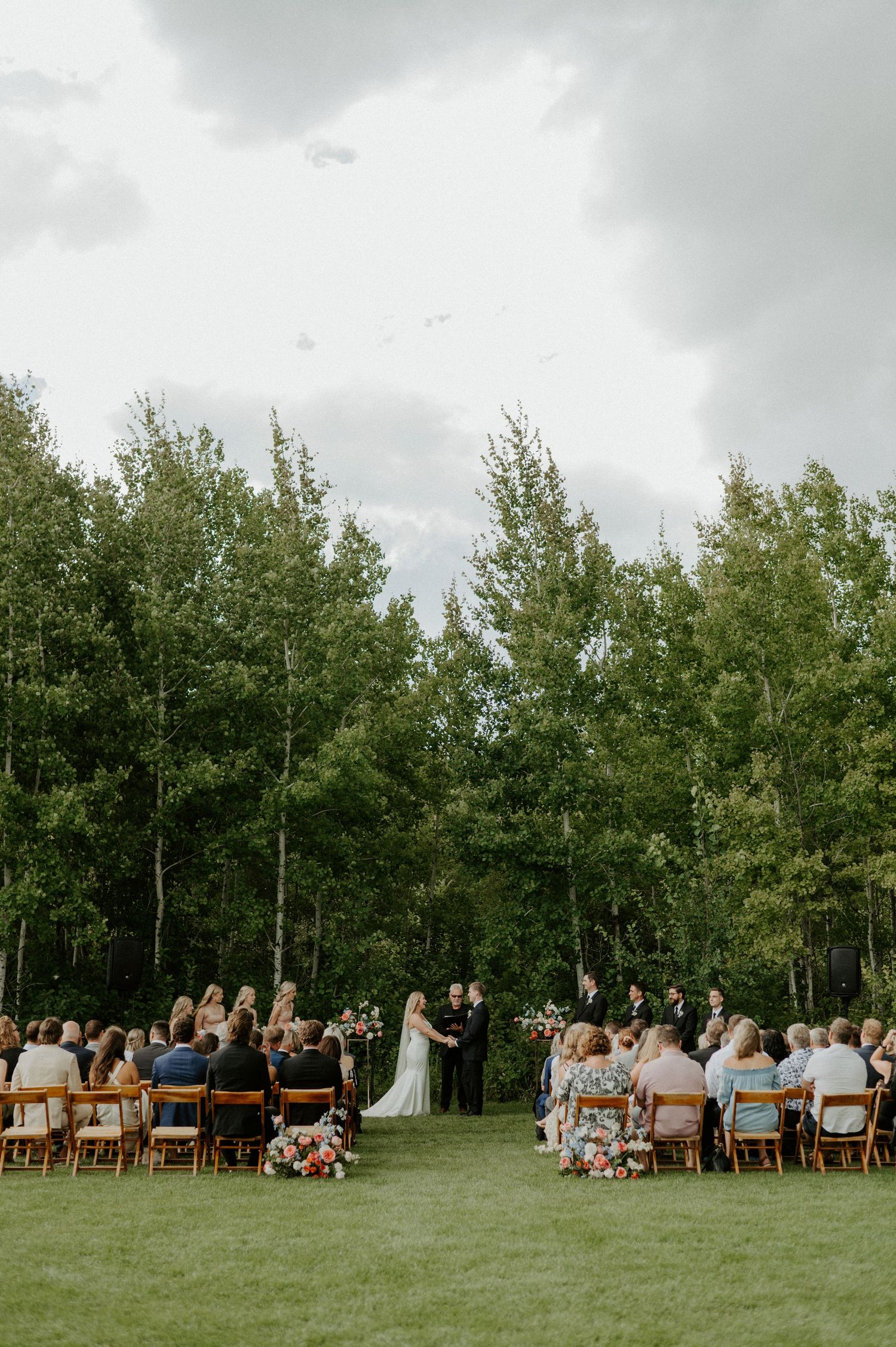 An outdoor wedding ceremony taking place on a green lawn surrounded by tall aspen trees under a cloudy sky.