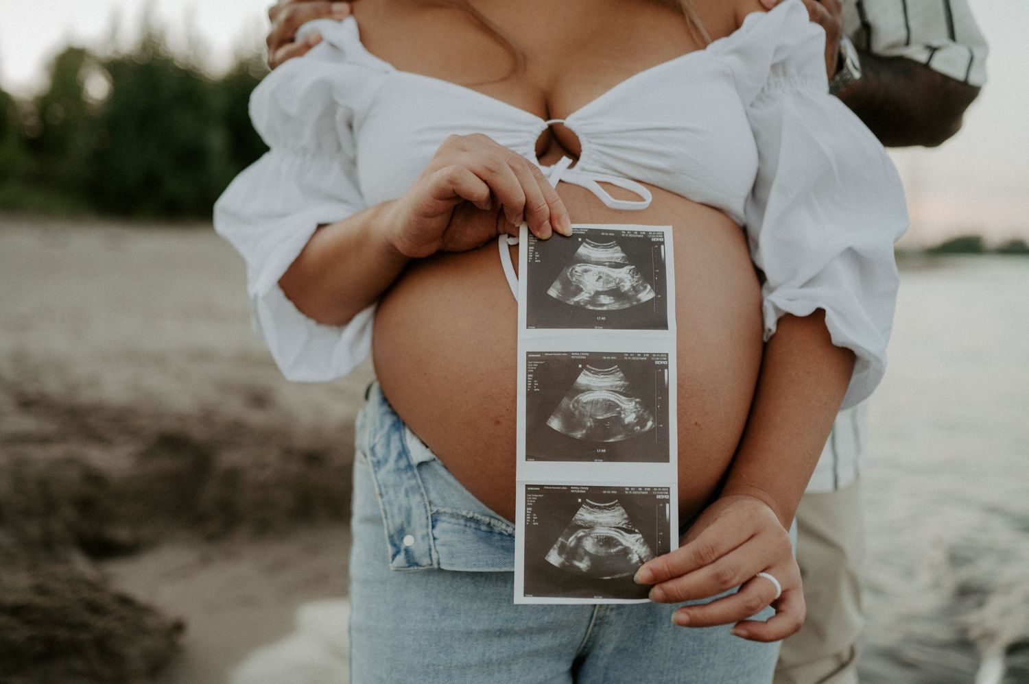Pregnant woman in white top holds ultrasound photos against her baby bump at sunset on beach.