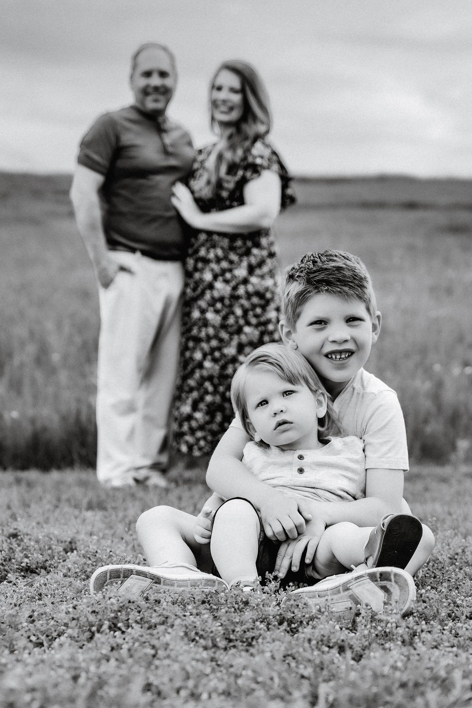 Black and white family portrait with two children sitting in grass foreground and parents embracing in blurred background.