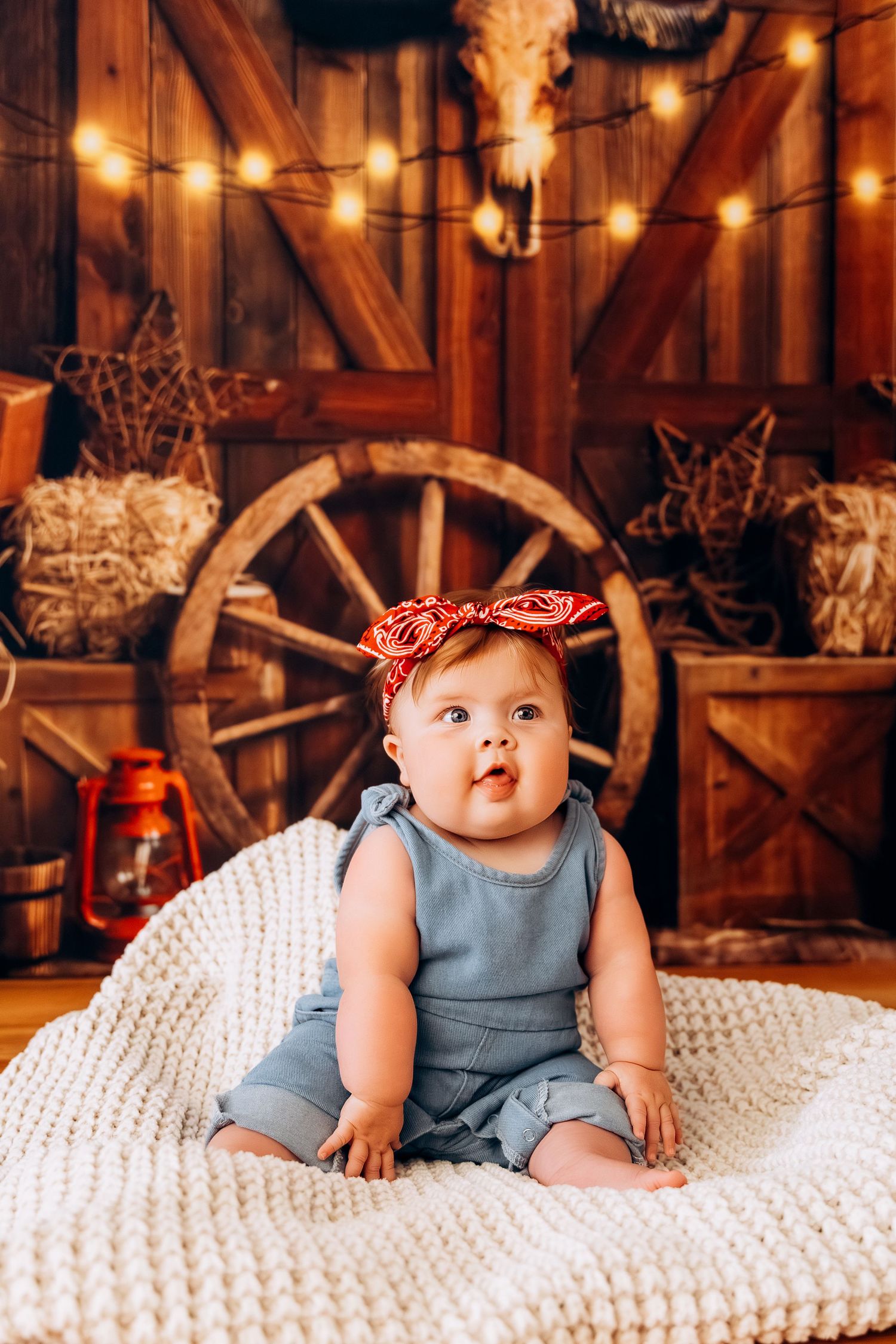 Cute baby girl in denim romper and red bow headband sitting on white blanket in rustic barn setting with string lights.