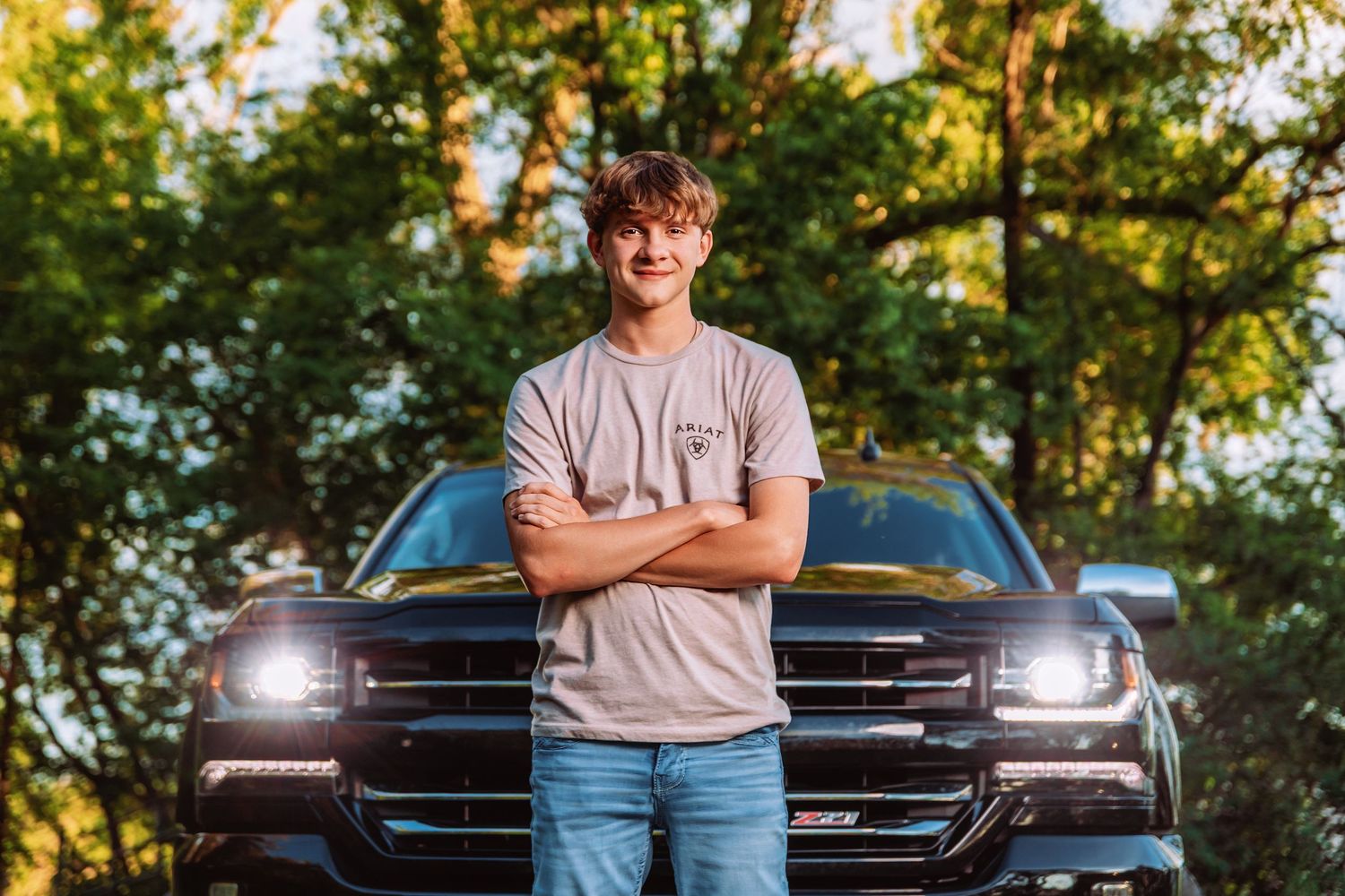 A person stands with crossed arms in front of a black pickup truck with illuminated headlights at sunset.